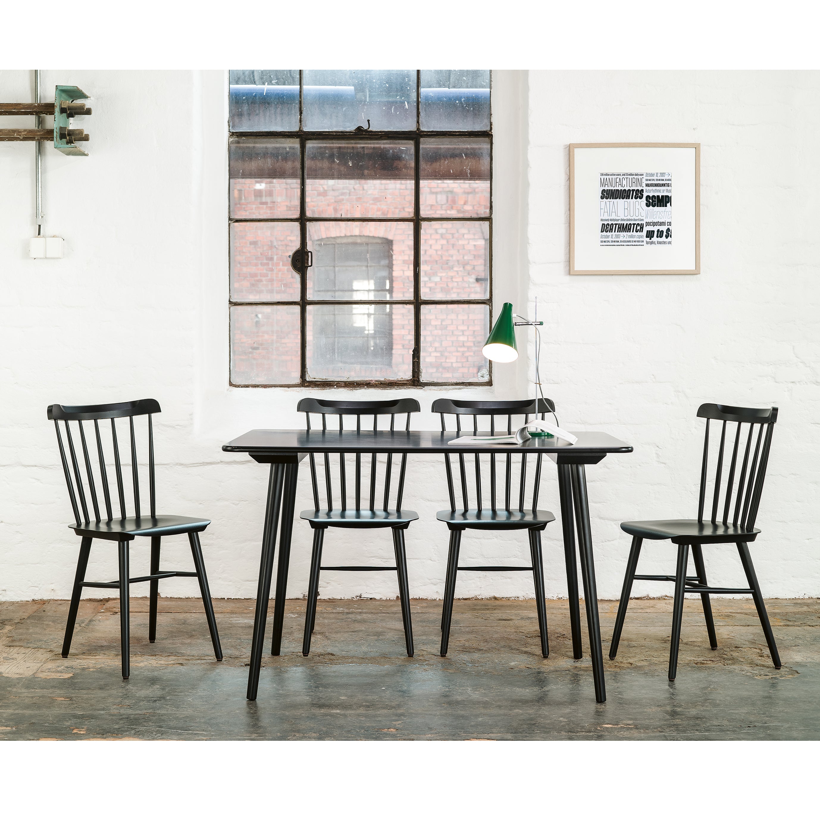 In a minimalist dining room, the retro-style black Table Ironica by TON pairs with four matching chairs. A green and white lamp sits on the solid beech table. A large window reveals a view of a brick building, while a framed typographic print adorns the white wall.