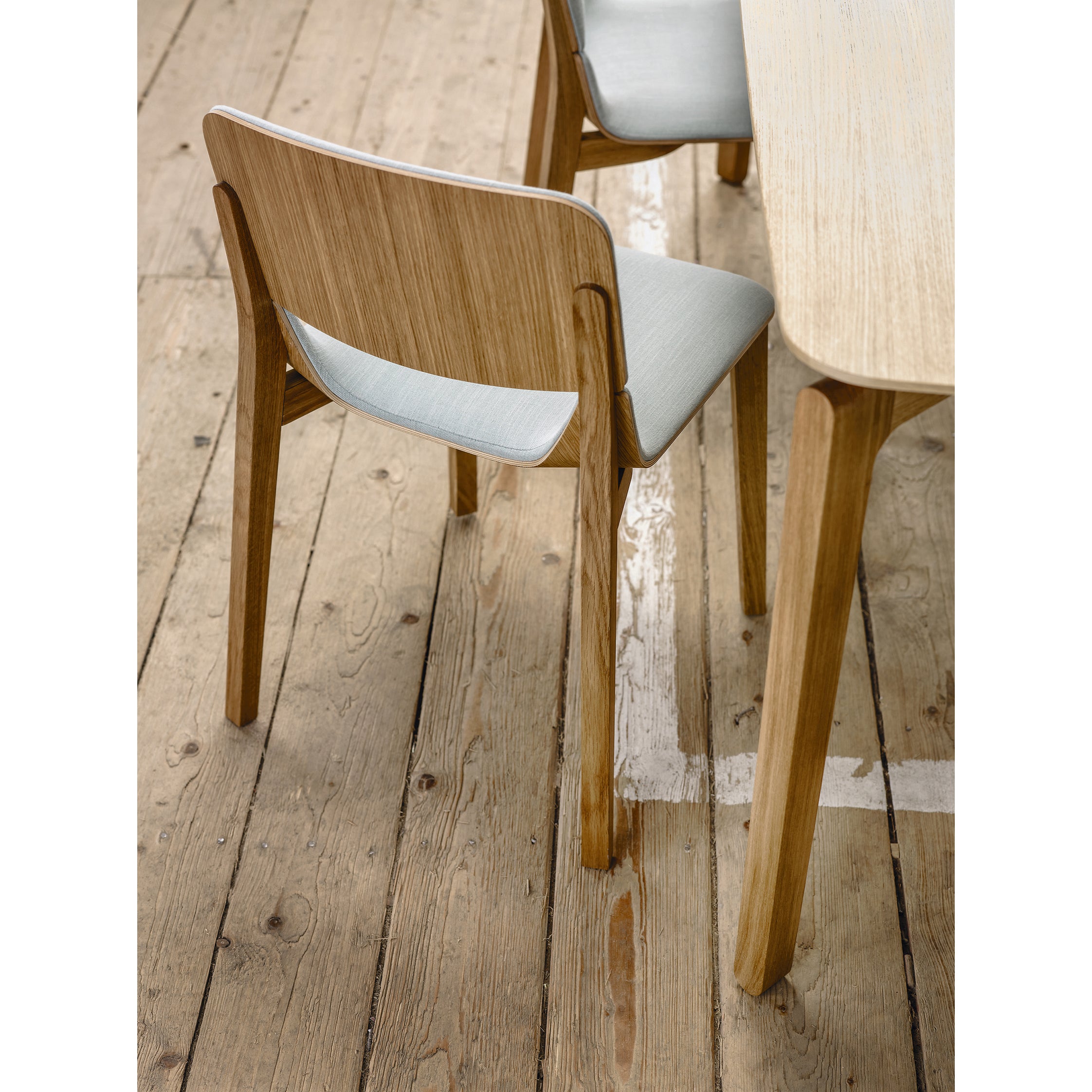 A Chair Leaf - Upholstered by TON with a light blue cushion is positioned next to a wooden table on a rustic floor, featuring visible planks and natural wear.