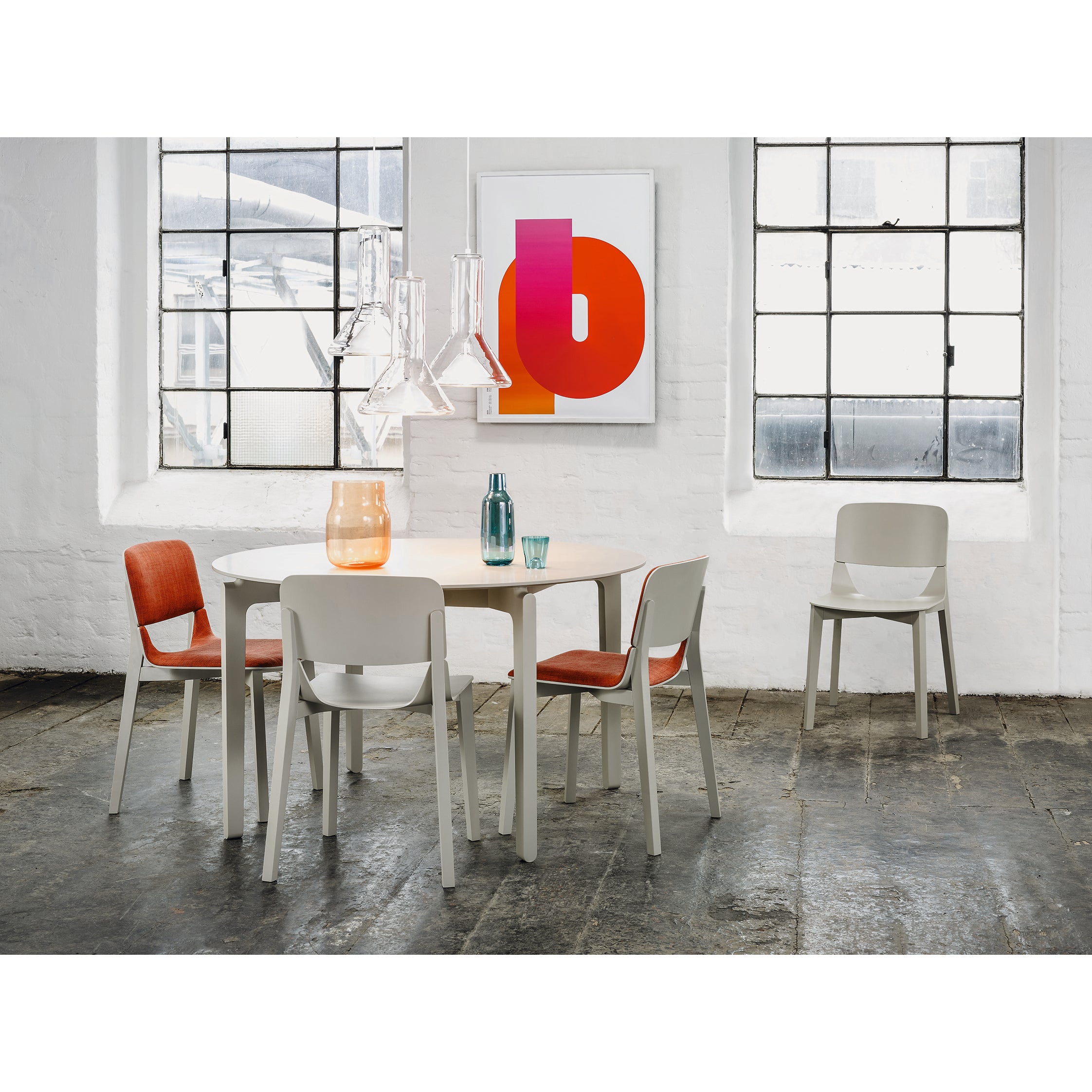 A modern dining area showcases a white table and four ergonomic TON Chair Leafs, two accented with orange cushions. A colorful abstract painting livens the white brick wall, while two industrial-style pendant lights and a window enhance the lively ambiance.