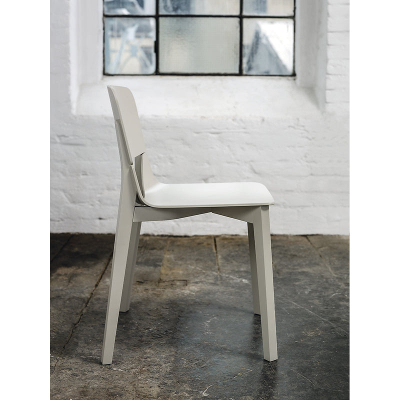 The TON Chair Leaf - Upholstered, a minimalist light gray dining accessory, sits on a rustic stone floor. A white brick wall with a partially frosted window behind it provides an industrial contrast to the modern design.