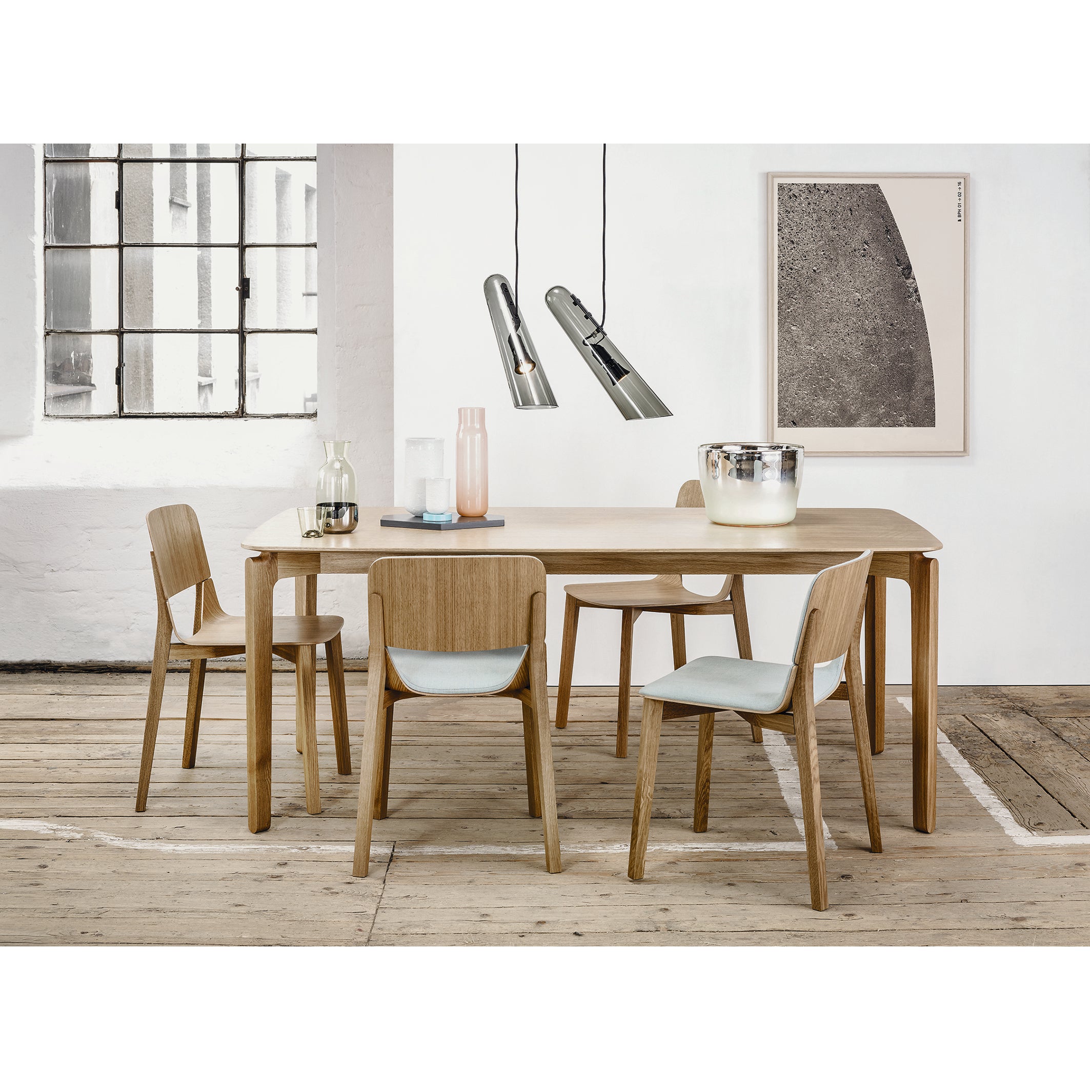 Modern dining room with a wooden table and four TON Chair Leaf dining chairs. Glassware and ceramics adorn the table. Pendant lights hang overhead, with a large window and abstract artwork decorating white walls, while the wooden floor enhances the stylish ambiance.