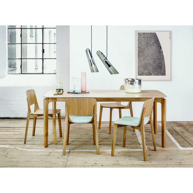 A modern dining room features a light-toned Leaf Table by TON with four matching ergonomic chairs. Two glass pendant lights hang above, while a large abstract artwork adorns the wall. Glass bottles and a metallic bowl adorn the tabletop on the wooden floor.