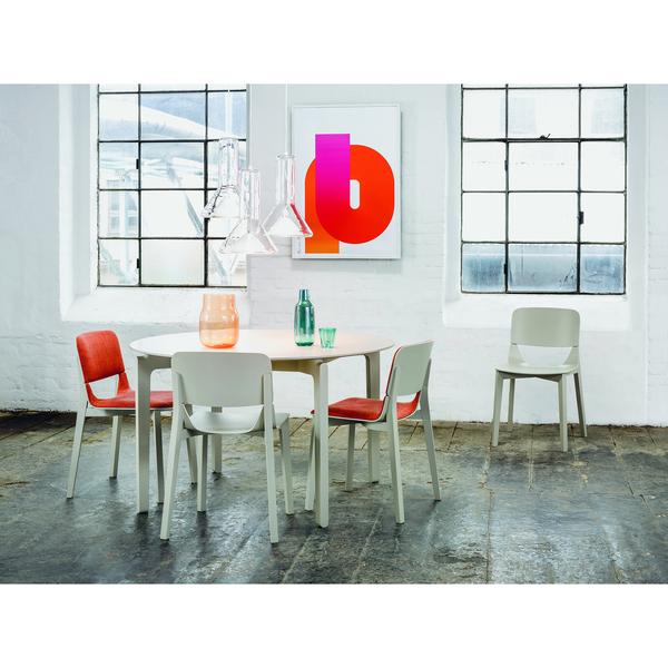 A modern dining room showcases a TON Leaf Table Round surrounded by five chairs, some with red cushions. A glass vase and green bottle decorate the table. A large abstract art piece graces the wall between two windows, completing this stylish space.