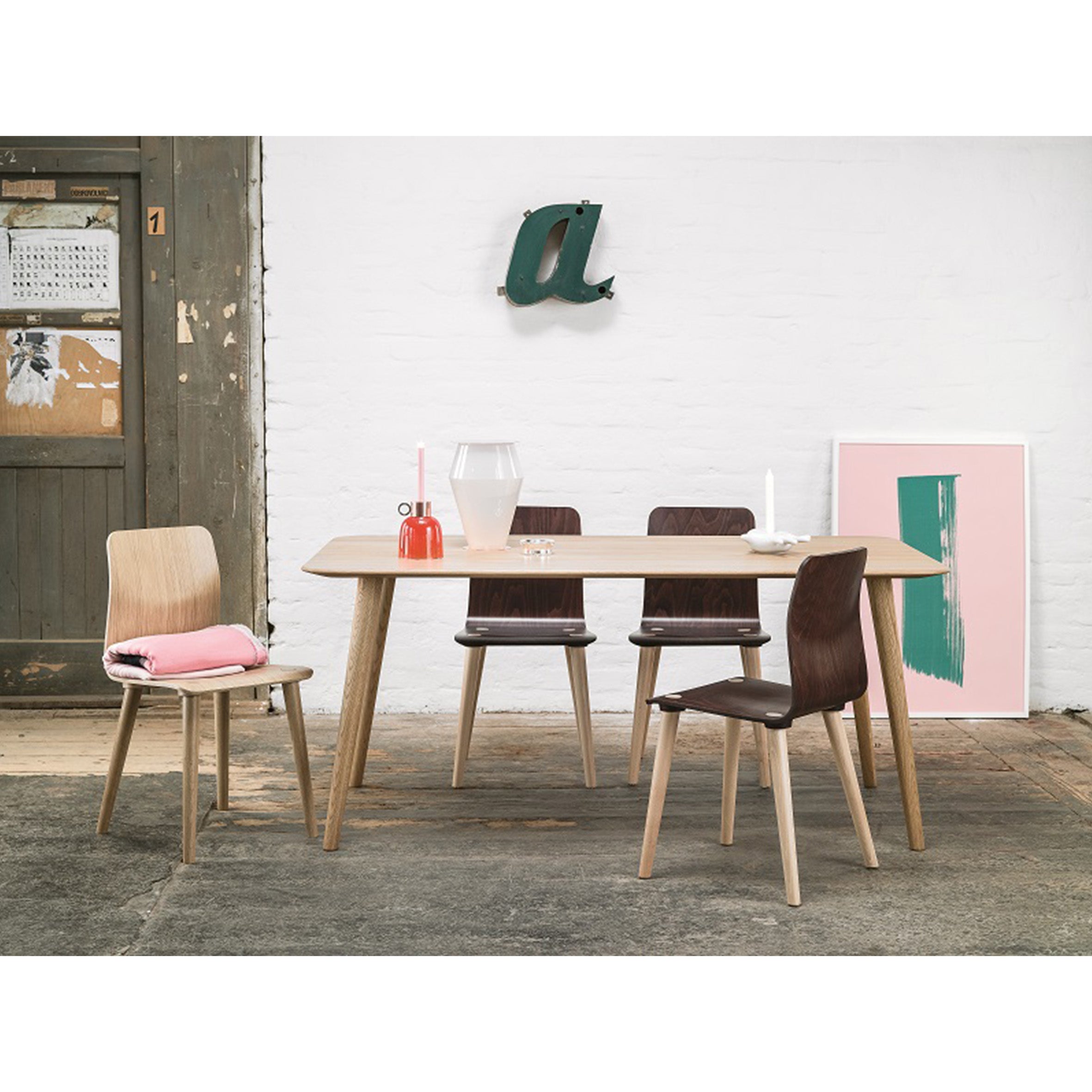 A simple dining room features a TON Table Malmö 706 with a wood tabletop, surrounded by dark wooden chairs, one accented with a pink cushion. On the table are a large jug and two candles. The wall displays artwork and a green a, while an ajar rustic door enhances the Scandinavian aesthetic.