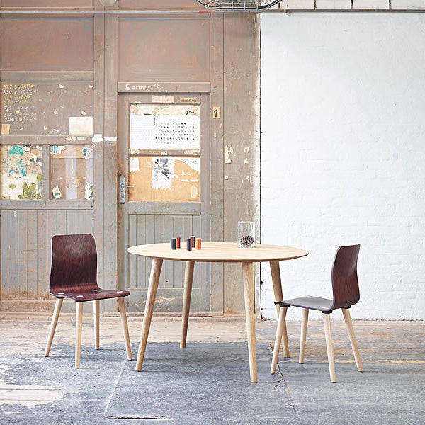 A minimalist room features a TON Table Malmö 707 with a round wooden top and two mismatched chairs. The rustic door with peeling posters contrasts against the white brick wall, adding a Scandinavian touch. A few small items adorn the tabletop.