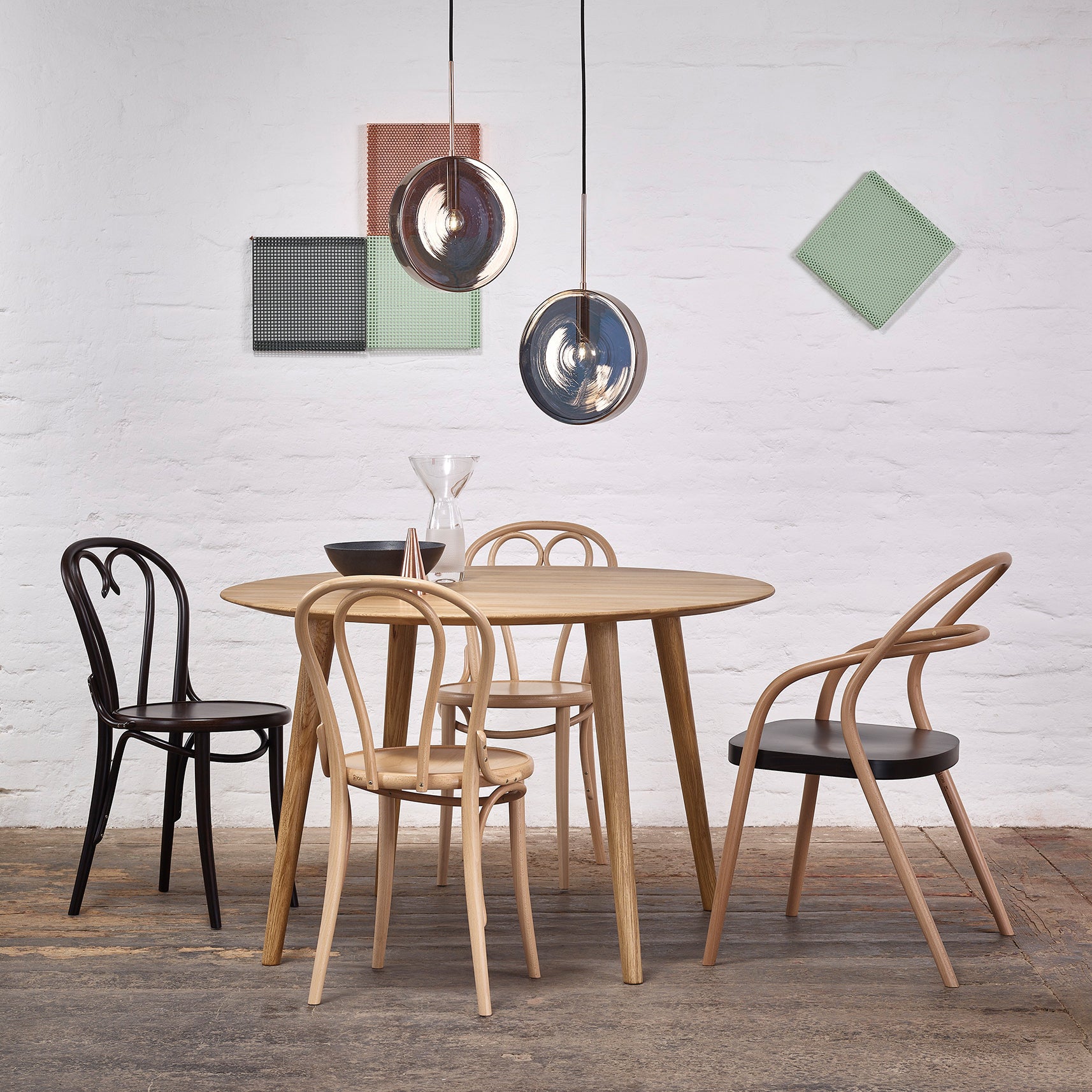 A modern dining space featuring a sleek TON Table Malmö 707, accompanied by four chairs—three in natural wood and one in black. Three stylish pendant lights illuminate the room, complemented by vibrant geometric wall art on a white brick wall. The wooden floor enhances its Scandinavian design appeal.