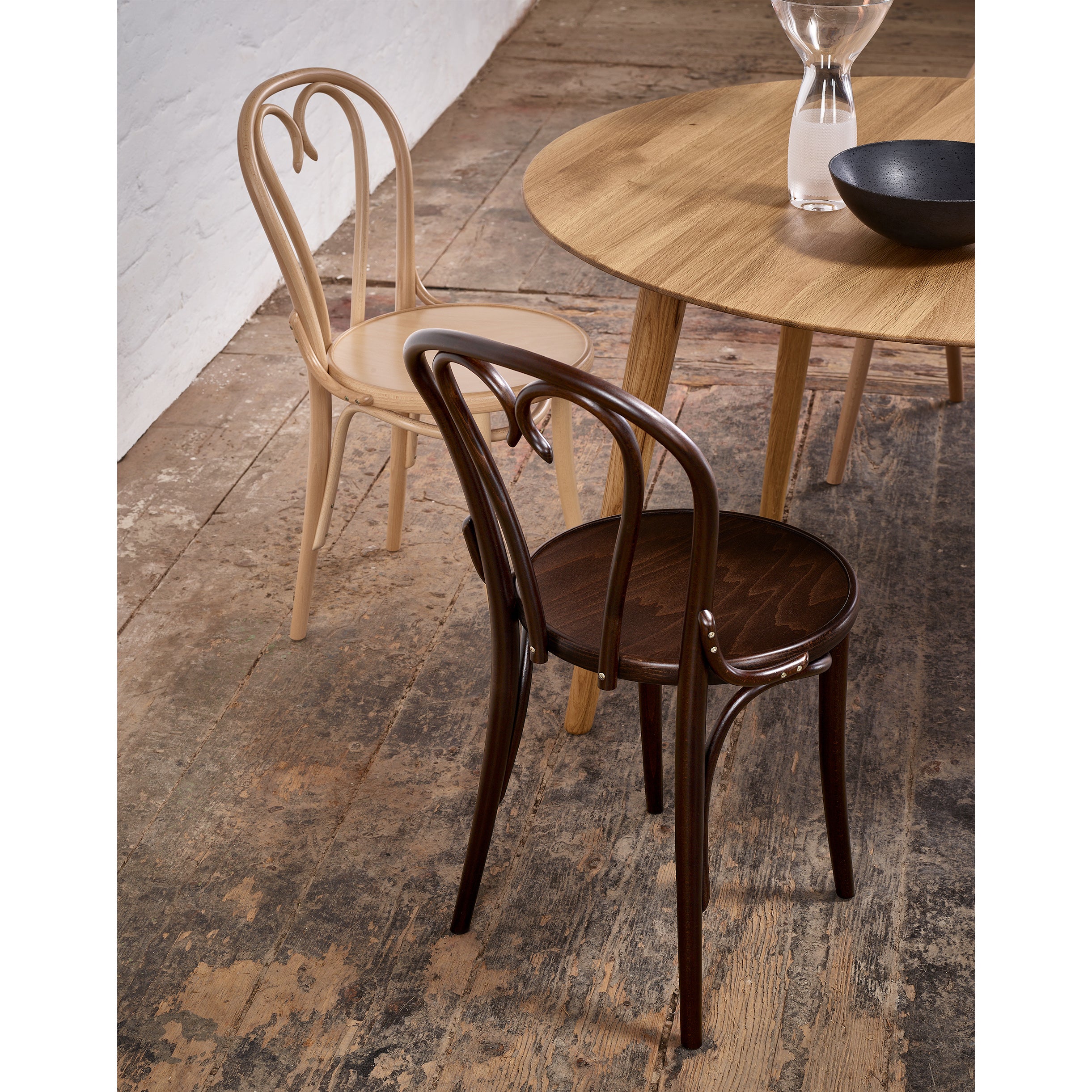 A light and dark curved wooden chair pair with a TONs Table Malmö 707 on a rustic wooden floor, accented by a glass vase and black bowl for a cozy Scandinavian design.