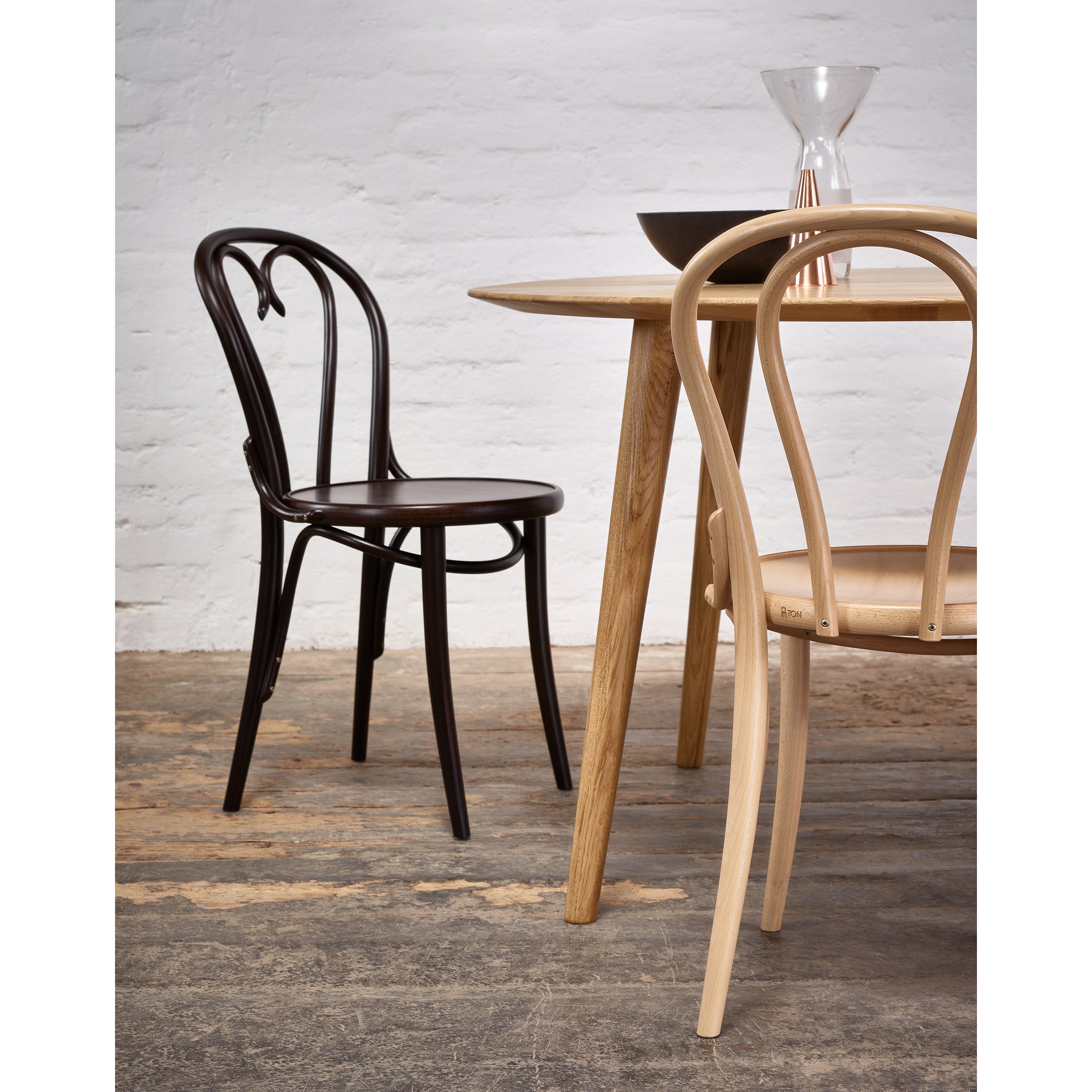 Two elegant wooden chairs, one dark and one light, surround a Table Malmö 707 by TON. The table features a bowl and a tall hourglass object, set against a textured white brick wall and rustic wooden floor, embodying the minimalist charm of Scandinavian design.