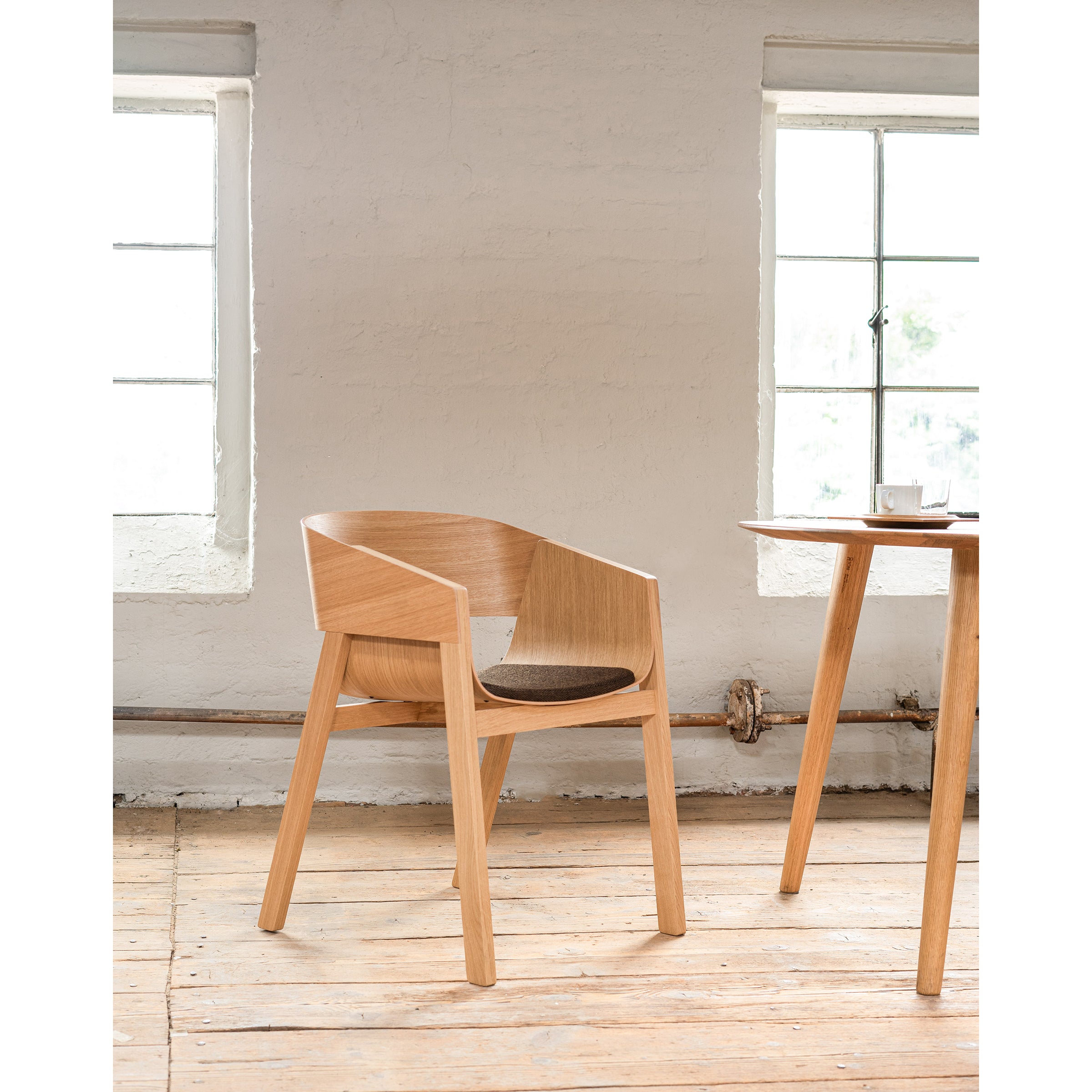 An Armchair Merano by TON, with a solid wood frame, curved backrest, and seat cushion, sits beside a wooden table. The bright room features two large windows, pristine white walls, and a beautifully finished wooden floor.