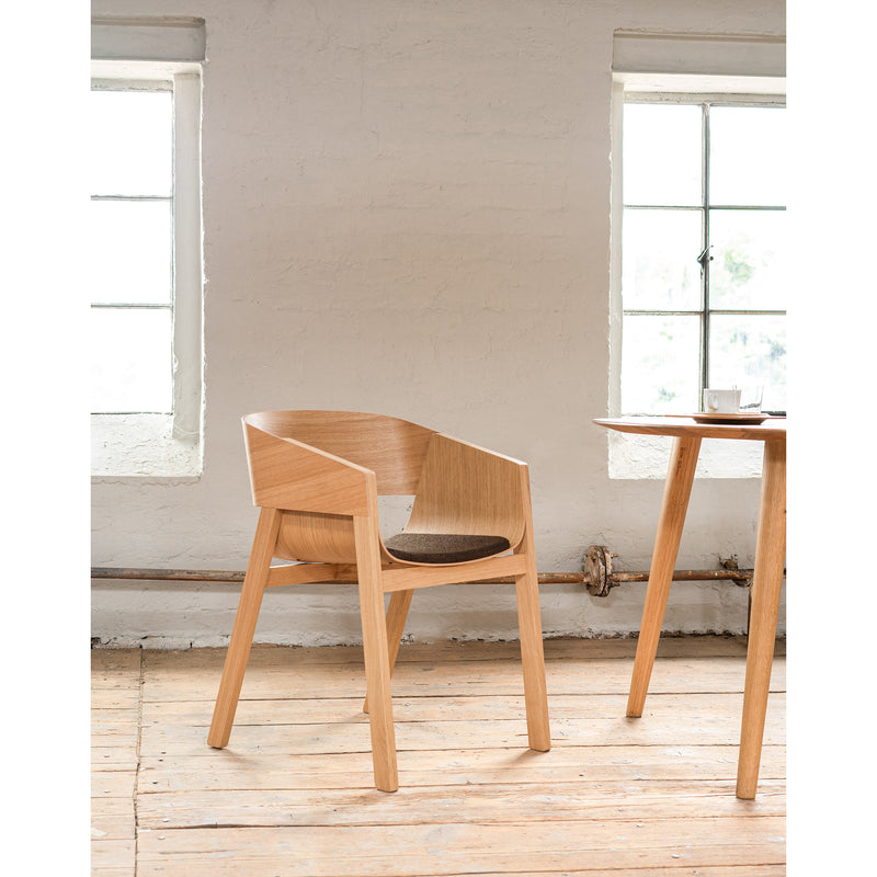 An Armchair Merano by TON, with a solid wood frame, curved backrest, and seat cushion, sits beside a wooden table. The bright room features two large windows, pristine white walls, and a beautifully finished wooden floor.