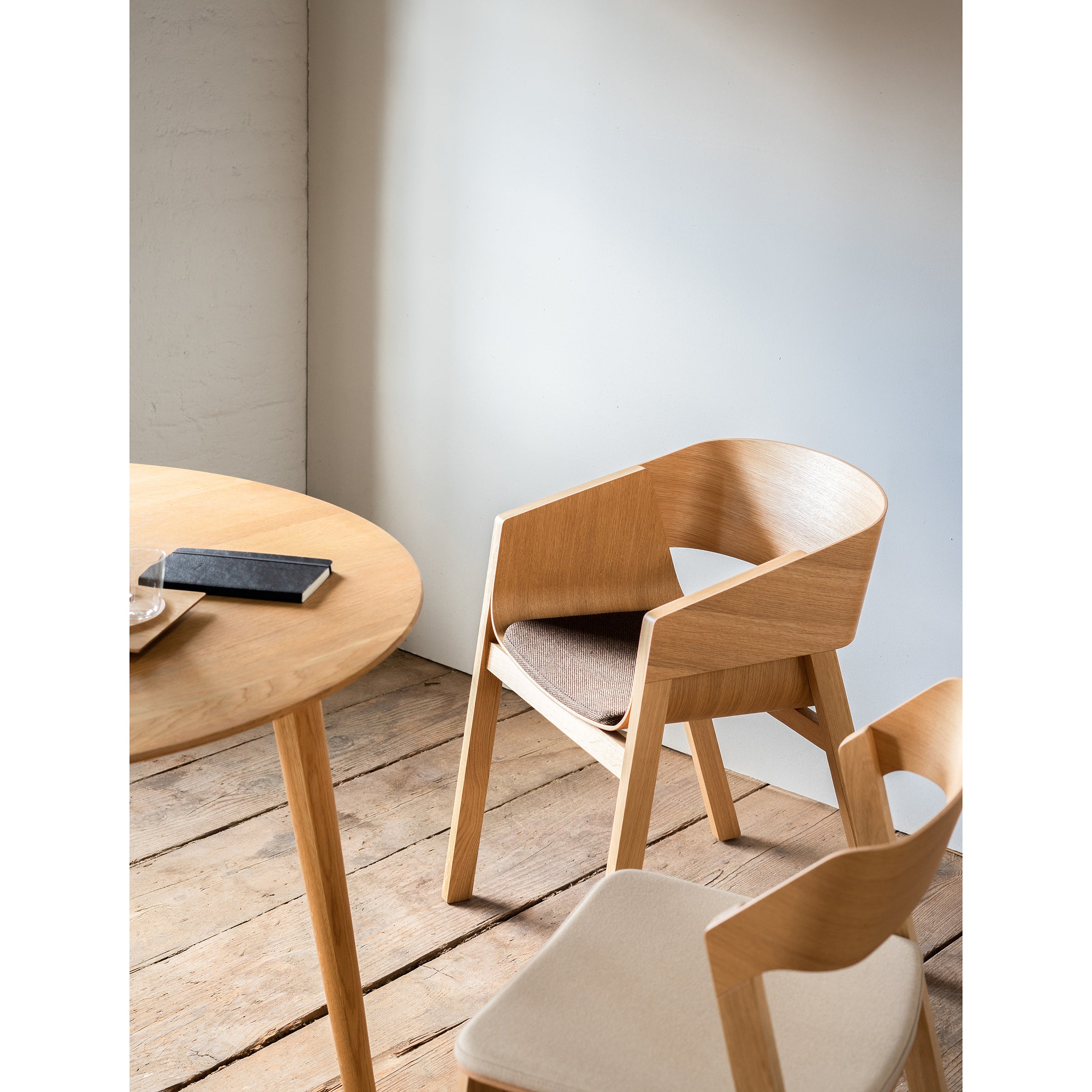 A minimalistic room features a round wooden table and two Armchair Merano by TON with light cushions. Their solid wood frames complement the notebook on the table, while natural light enhances the wooden flooring, creating a serene ambiance.