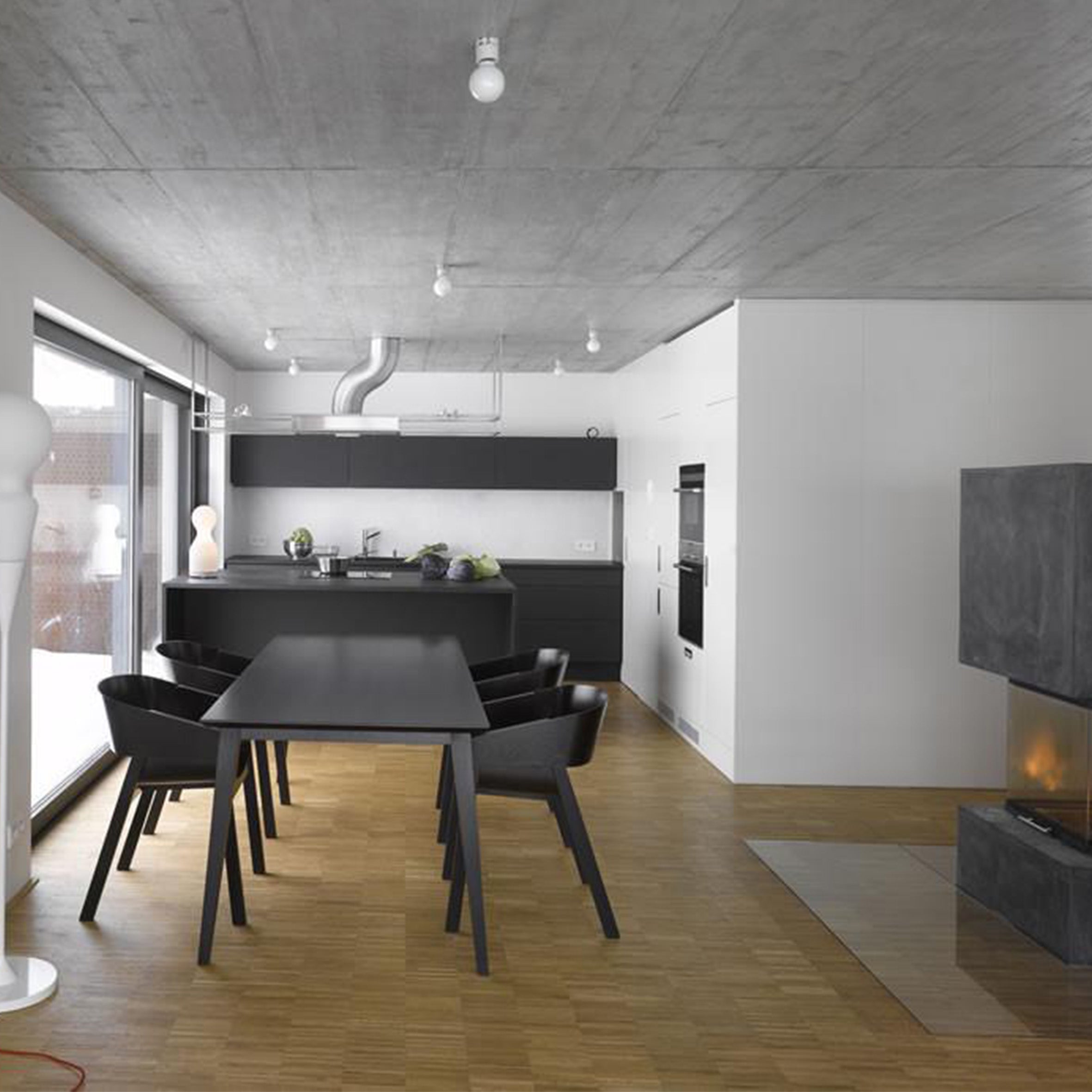 Modern kitchen and dining area in a minimalist design with a black dining table and chairs, sleek cabinetry, parquet floor, wall-mounted oven, large window, and fireplace. The TON Armchair Merano with plywood shells complements the industrial vibe of the concrete ceiling.