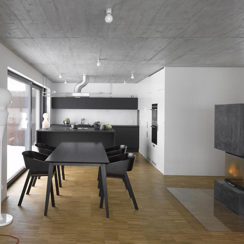 Modern kitchen and dining area in a minimalist design with a black dining table and chairs, sleek cabinetry, parquet floor, wall-mounted oven, large window, and fireplace. The TON Armchair Merano with plywood shells complements the industrial vibe of the concrete ceiling.