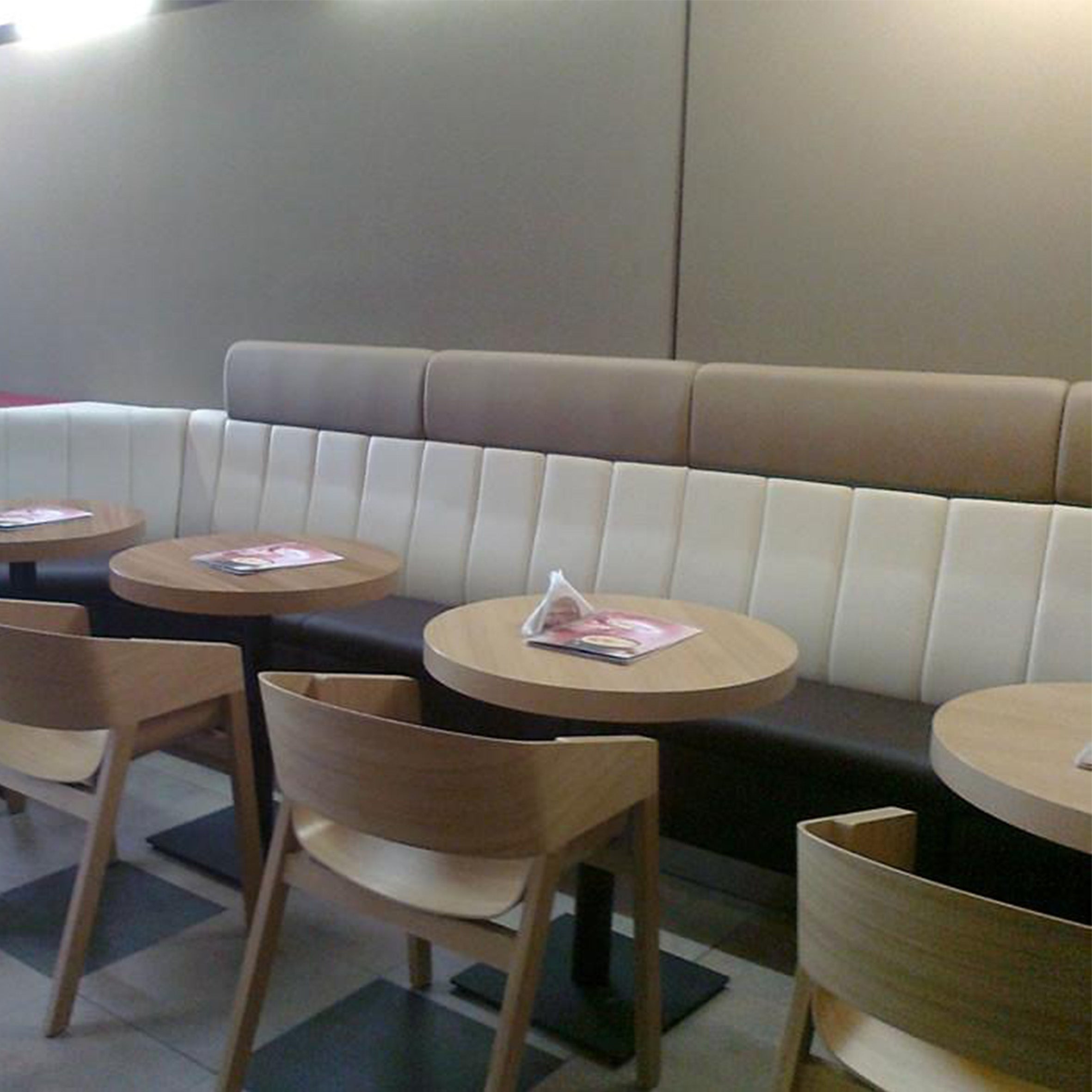 The modern cafe interior showcases beige and brown padded benches along the wall. Solid wood frame chairs accompany round tables topped with magazines, while Armchair Merano by TON adds elegance to the softly lit, plain walls.