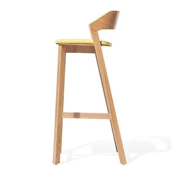 The Merano barstool by TON showcases a minimalist side view with a solid wood frame, yellow cushioned seat, and slightly angled backrest. Its clean lines and natural finish embody modern simplicity.