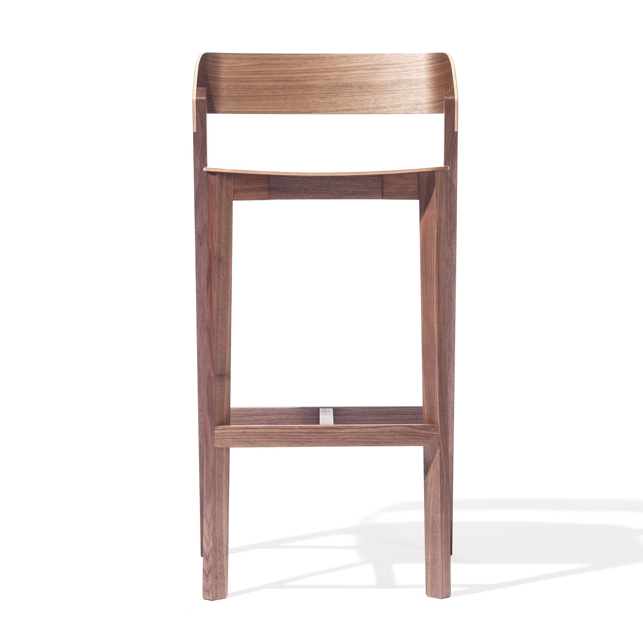 The Barstool Merano by TON showcases a minimalist design with a rectangular seat, low backrest, and straight solid wood legs connected by a footrest bar, all against a plain white background.