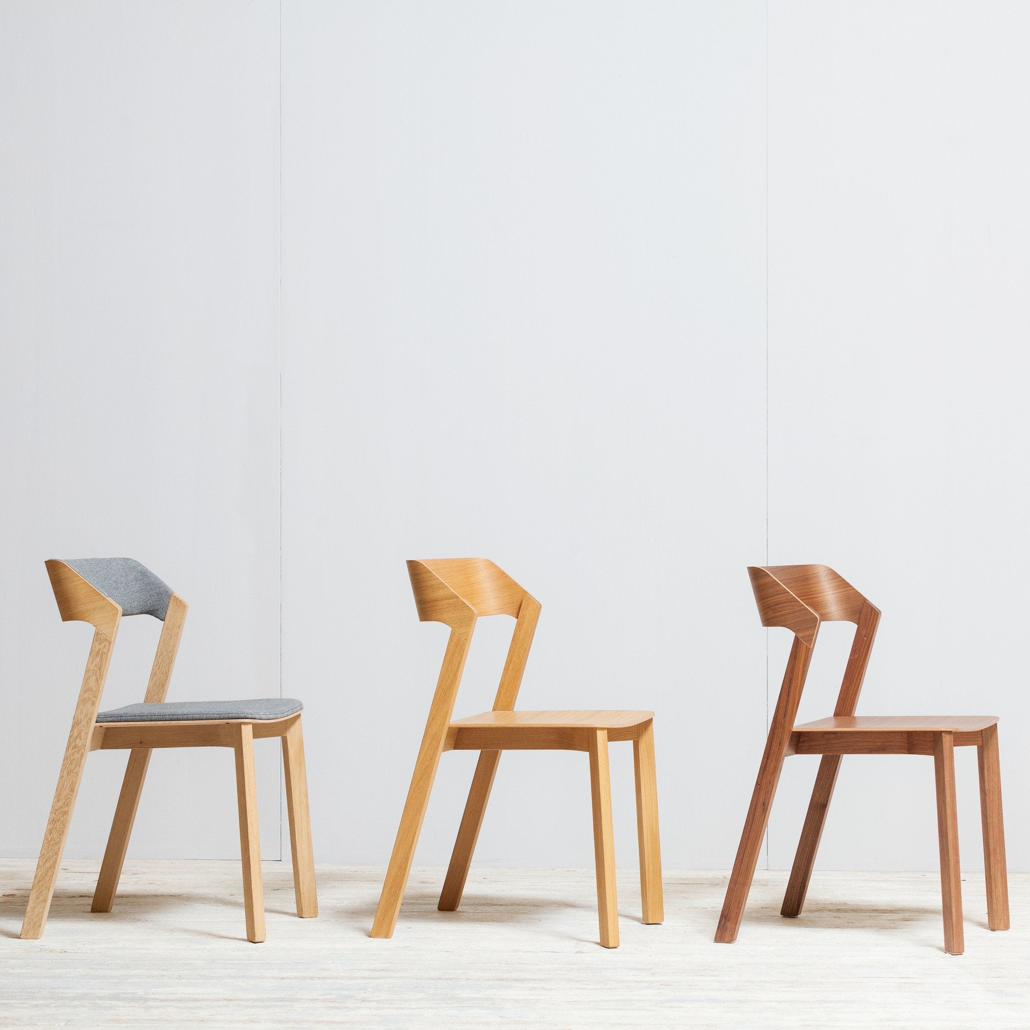 Three TON Chair Merano wooden chairs, each with distinct grain and tone, resemble sleek dining pieces. The first features a gray cushion; all have angular, minimalist designs. Crafted from solid wood, they rest on a light floor against a plain white wall.