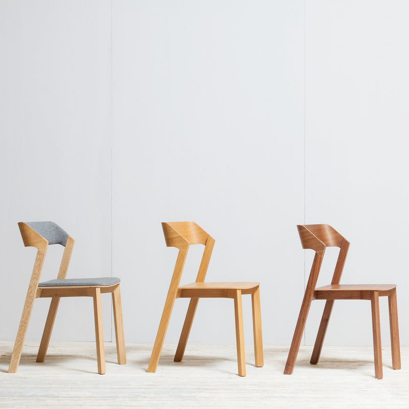 Three modern chairs, all showcasing the elegance of the Merano chair, are lined up against a white wall. The first chair features bent plywood with a gray cushion, produced by TON and is currently on sale. The second is light wood and the third is dark wood; all have angled backrests in minimalist designs.