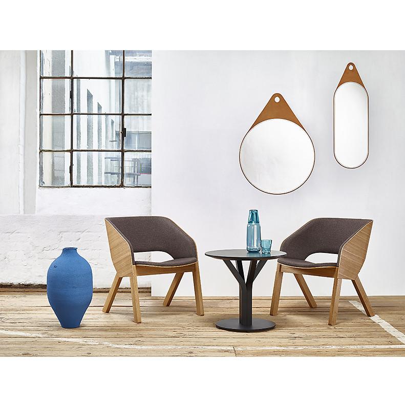 In this minimalist room, two TON Armchair Merano Lounges with solid wood legs flank a small round table adorned with a blue vase and water carafe with glasses. Above, two wall mirrors reflect the large windows in the background.