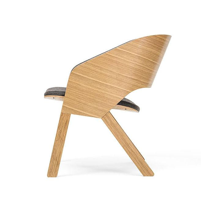 The Armchair Merano Lounge by TON features a sleek, curved wooden backrest and bent plywood seat. Its innovative minimalist design includes solid wood legs with a simple front leg and slanted back leg, while the seat is upholstered in dark fabric.