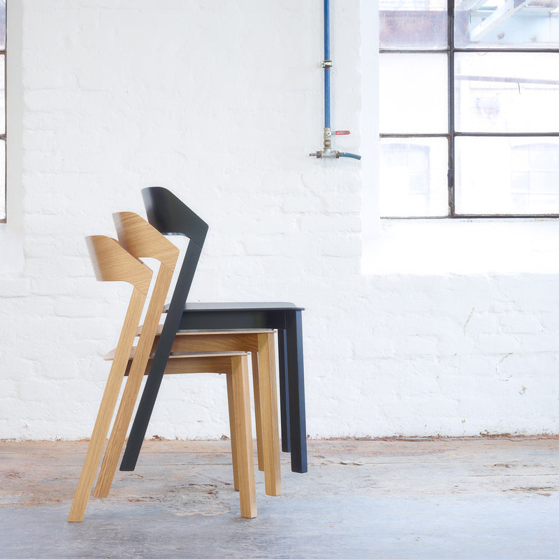 Three upholstered Merano chairs by TON, elegantly stacked, enhance a minimalist, light-filled room. White brick walls and a large window complement the visible blue pipe, while the concrete floor adds to the industrial aesthetic.