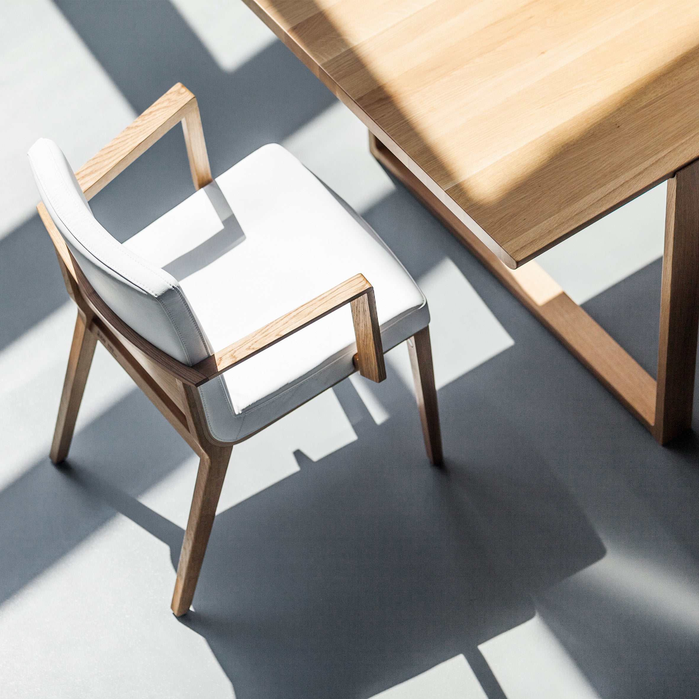 The Armchair Moritz, from the TON collection by Kai Stania, has white cushions and wooden arms and legs. It pairs beautifully with a light wooden table, as sunlight creates sharp shadows on the gray floor, highlighting their minimalist and modern design.