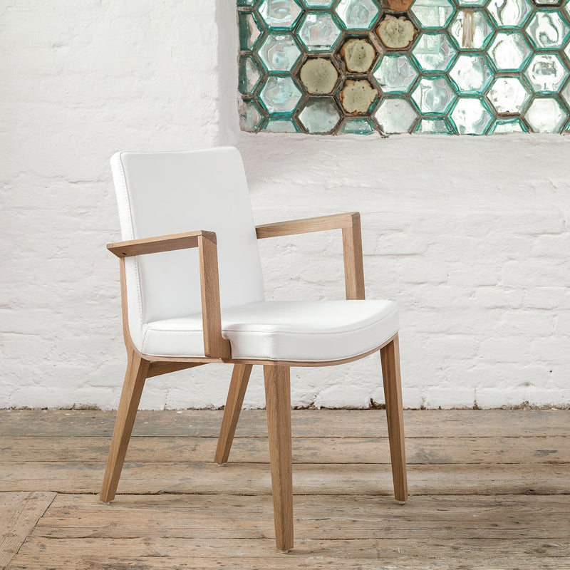 The Armchair Moritz by TON, designed by Kai Stania, features wooden legs, a white cushioned seat and back with armrests. It complements a wooden floor next to a white brick wall adorned with hexagonal glass-paned decorative window.