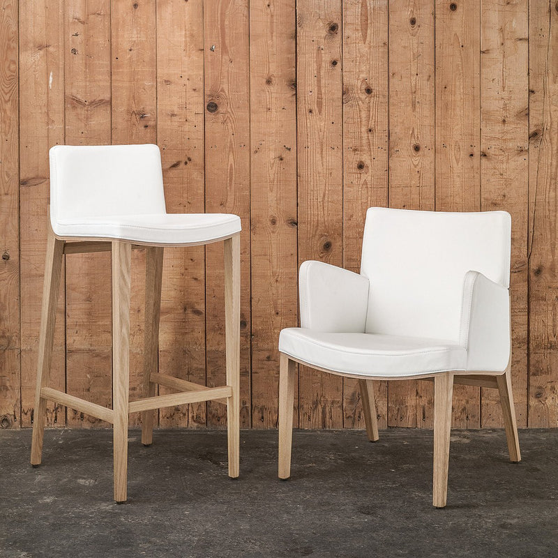 The white Barstool Moritz by TON and an armchair with wooden legs are set against a rustic wooden wall. A concrete floor adds to the minimalistic and cozy vibe, highlighting premium ergonomic furniture design.