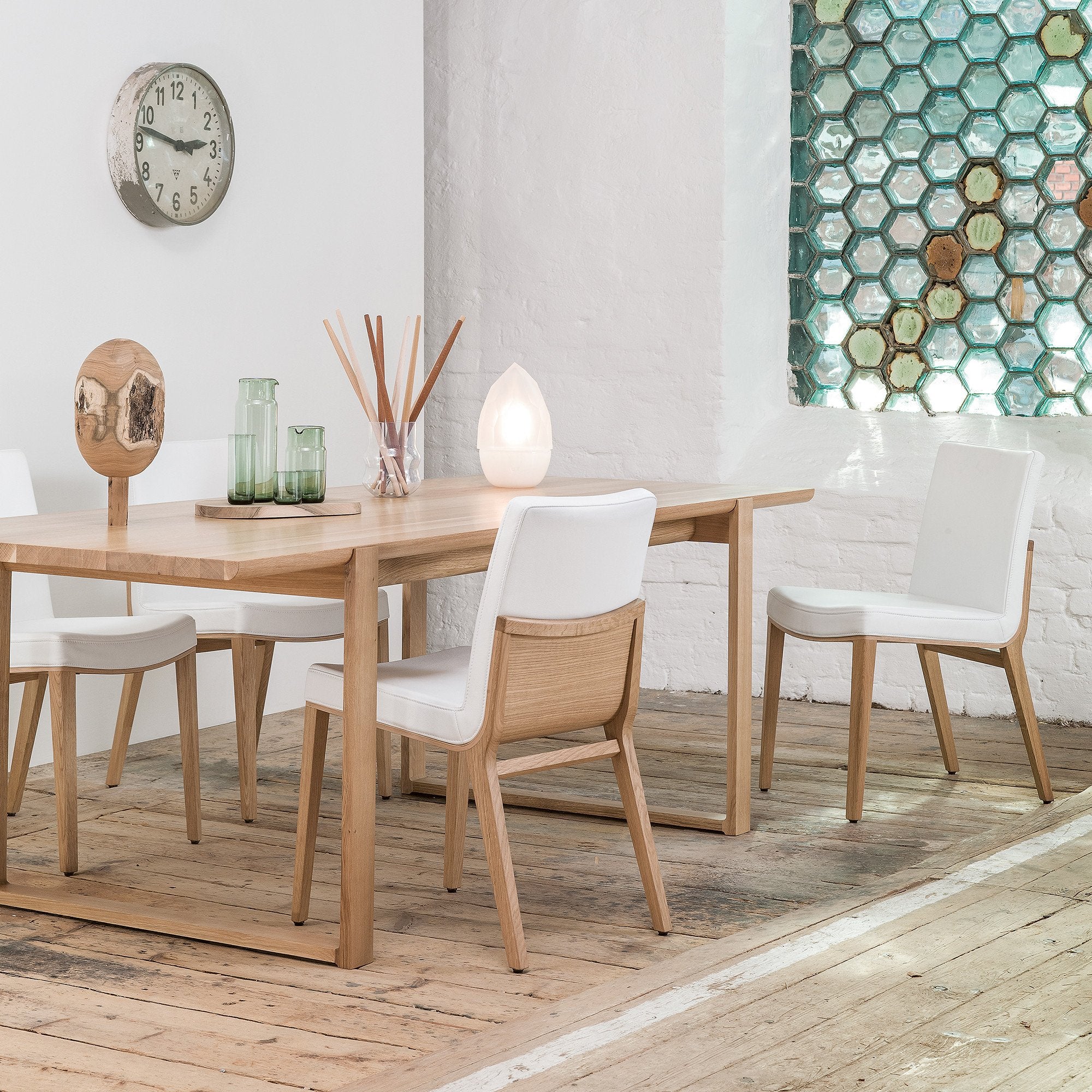 A modern dining room showcases a wooden table, several chairs, and premium white-cushioned seats. A clock hangs near a hexagonal stained glass window. The glowing lamp by TONDesigner adds a minimalist yet cozy atmosphere, complemented by the Moritz chairs from TON.