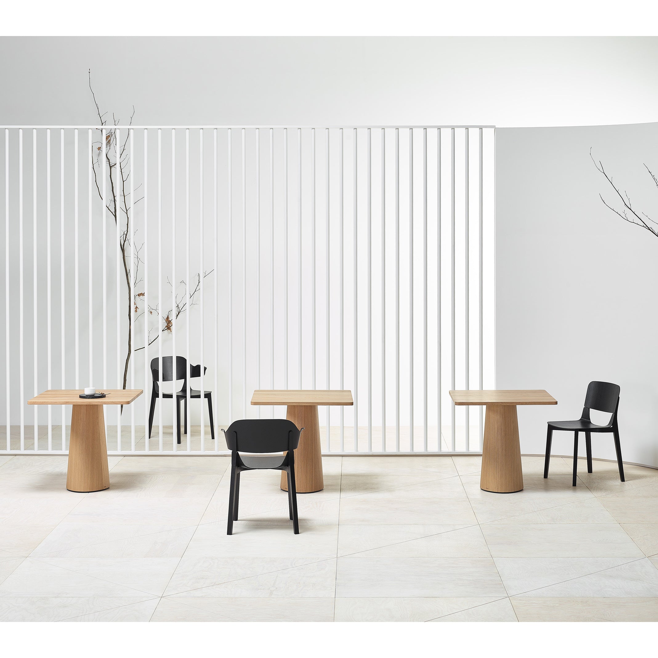 A minimalist room showcases three P.O.V. Table 460 pedestal tables by TON with organically shaped bases, paired with three black chairs. Bare branches accentuate a white slatted wall in the background, creating a serene, modern vibe.