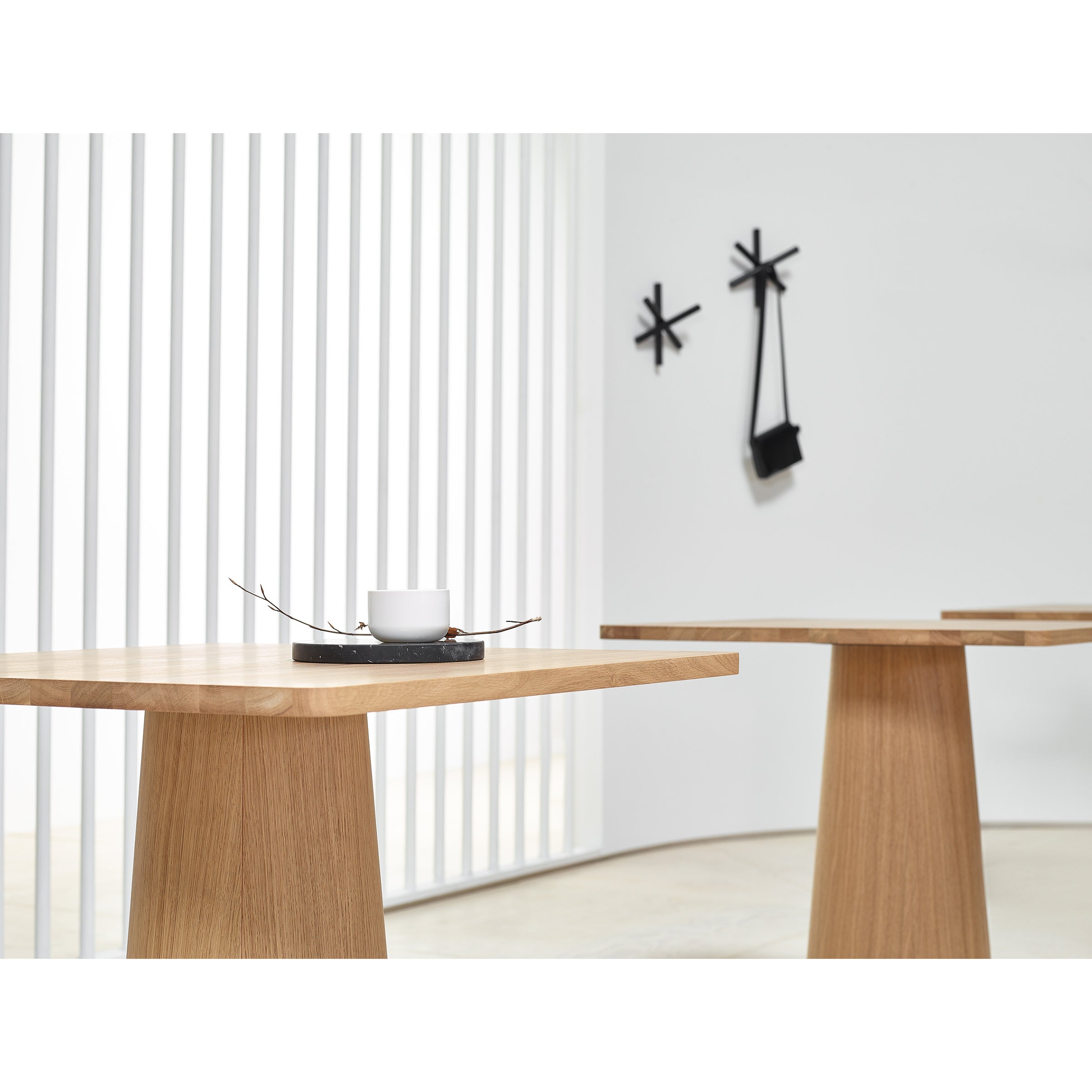 Minimalist interior with two P.O.V. Table 460 tables by TON, featuring organic bases. Each table holds a black tray with a white cup. In the background are white vertical blinds and a modern black clock without numbers.