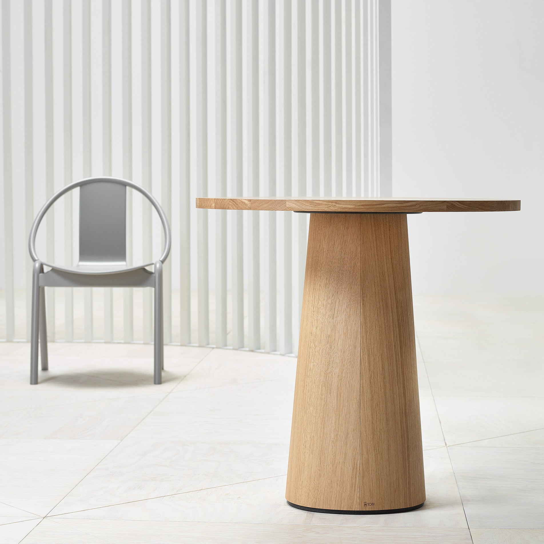 A modern setting highlights the P.O.V. Table 461 by TON with an organic-shaped base upfront and a sleek gray metal chair featuring a circular backrest behind. Light floors and walls harmonize seamlessly with vertical slats in the backdrop.