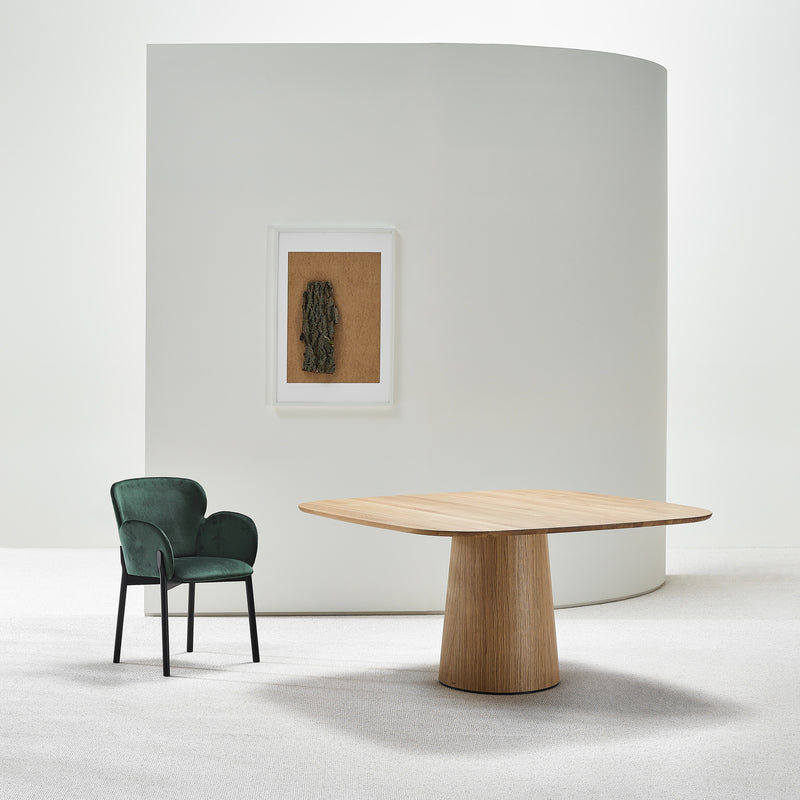 A modern interior features a green velvet chair beside the TON P.O.V. Table 462 with an organically shaped base and square top. A framed abstract artwork hangs on a curved light grey wall, creating harmony, while soft white carpet covers the floor.