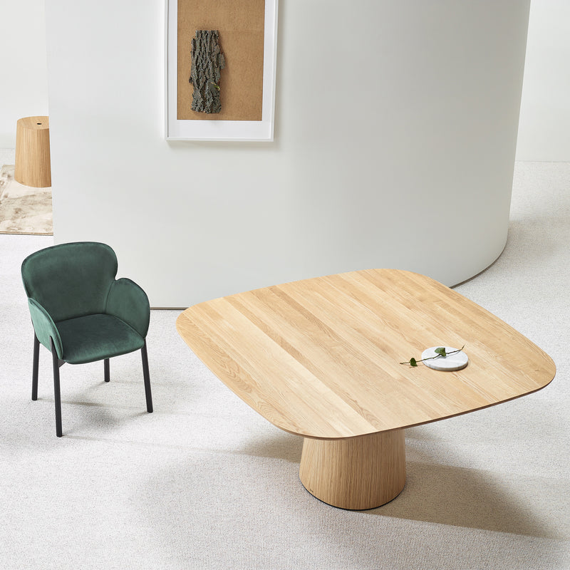 The modern room showcases a P.O.V. Table 462 by TON with a square wooden top and organically shaped base, accompanied by a green velvet chair with black metal legs and abstract wall art. Minimalistic decor features neutral-toned walls and carpet, embodying modular design principles.