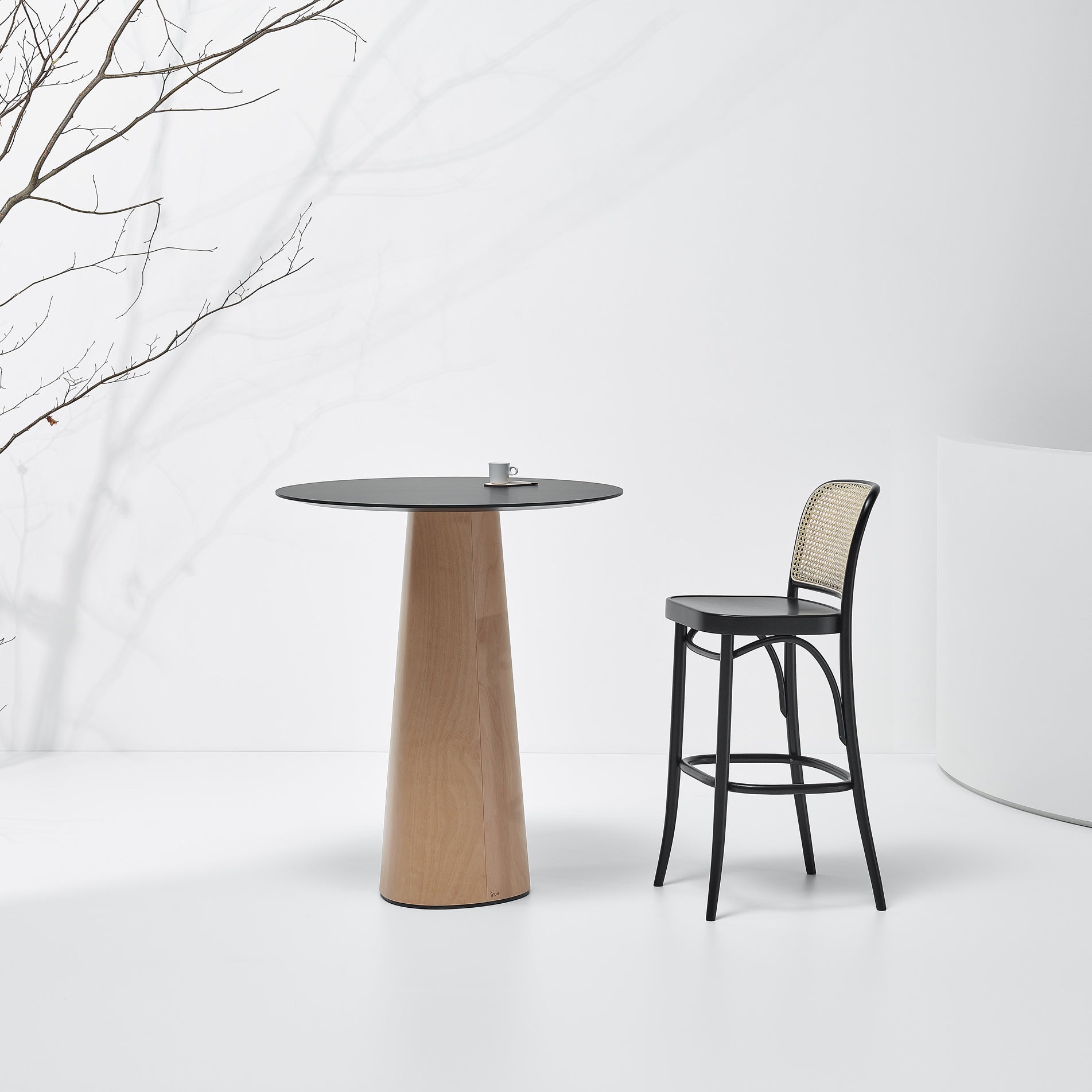 A minimalist scene features the P.O.V. Table 463 by TON with a solid wood tabletop and a black bar stool with a woven backrest. A small cup rests on the table, and a bare branch extends from the left against the pristine white background.