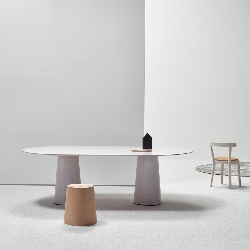 A minimalist room showcases a TON P.O.V. Table 464 with a long oval top and uniquely shaped base, flanked by two wooden stools on a light gray carpet. A simple white chair sits near a circular rug in the corner, with light gray walls enhancing the serene atmosphere.