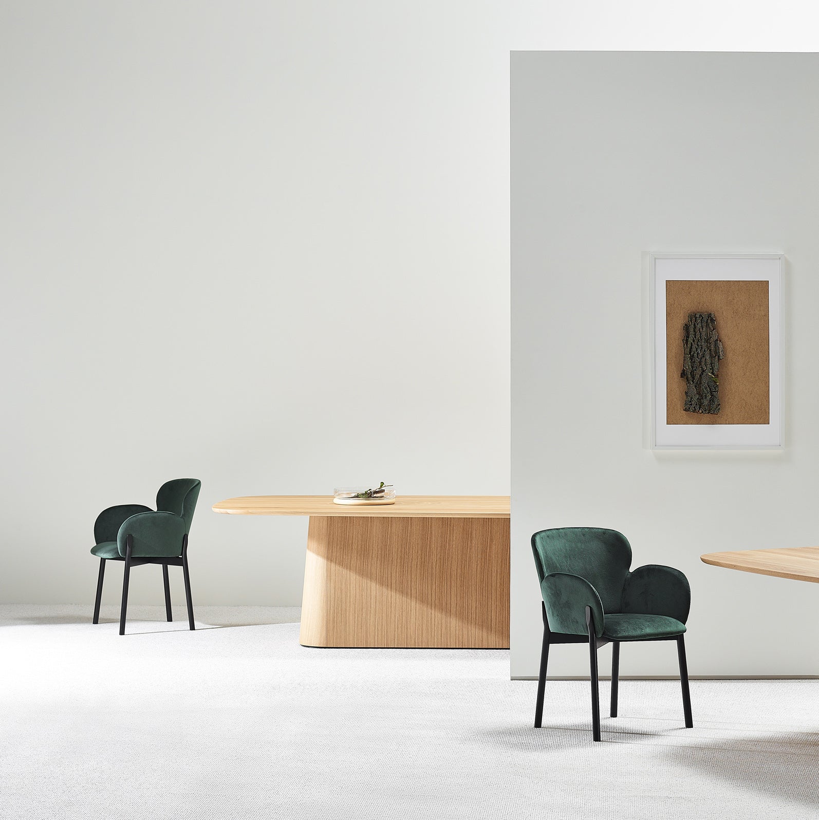 The minimalist room features a solid wood P.O.V. Table 466 by TON, paired with two green upholstered chairs and framed artwork. A light wooden panel divides the space, showcasing a simple, modern design that highlights an elegant aesthetic through its modular elements.