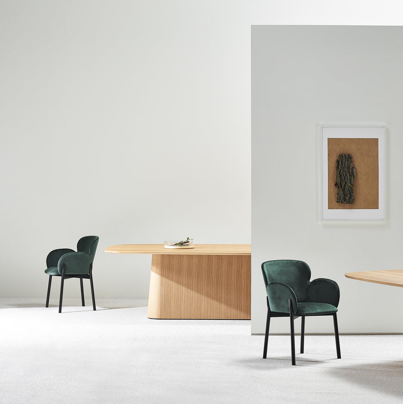 The minimalist room features a solid wood P.O.V. Table 466 by TON, paired with two green upholstered chairs and framed artwork. A light wooden panel divides the space, showcasing a simple, modern design that highlights an elegant aesthetic through its modular elements.