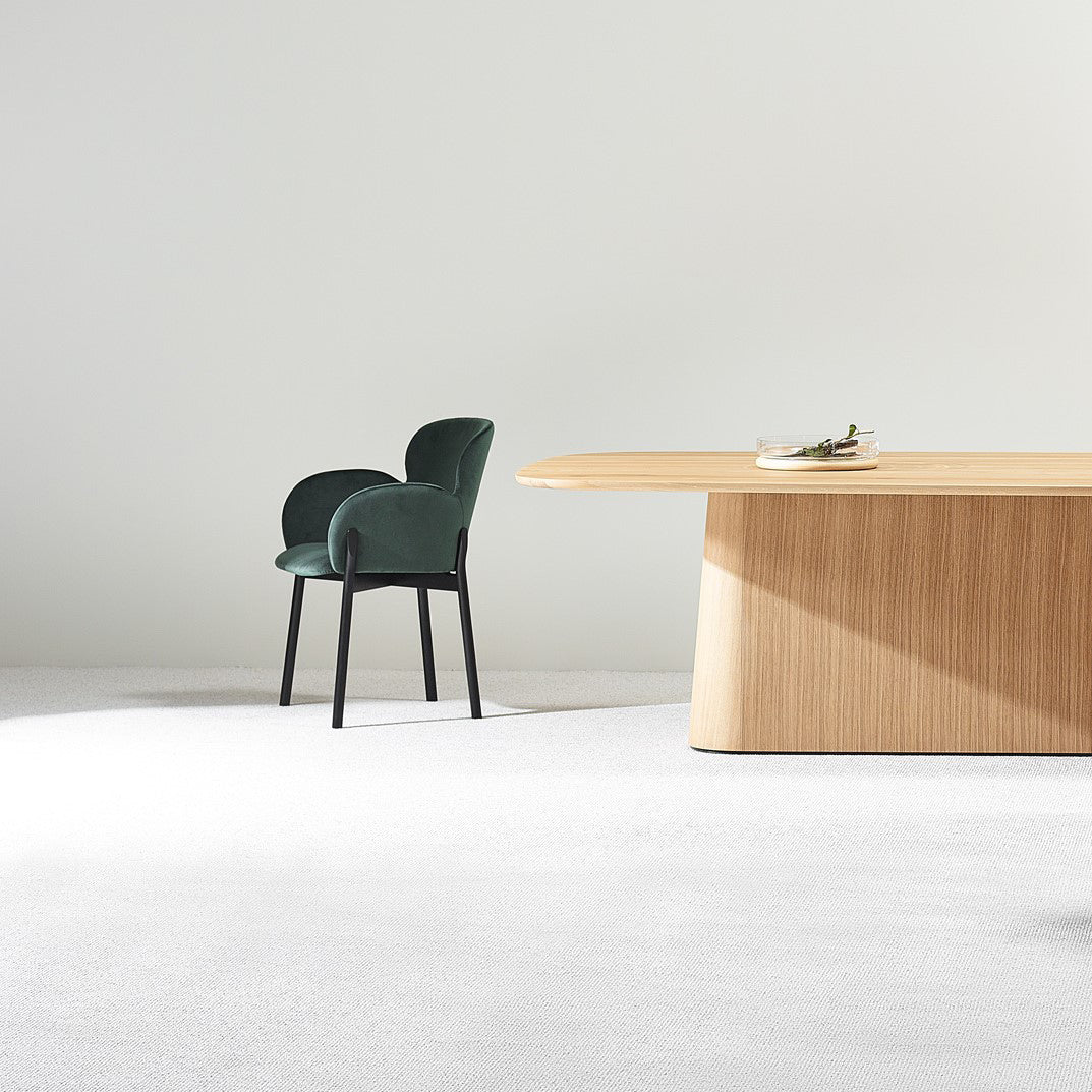 A minimalist scene showcases a green upholstered chair and the P.O.V. Table 466 by TON against a light gray wall and carpeted floor, with a small book and glasses on the table illuminated by soft natural light.