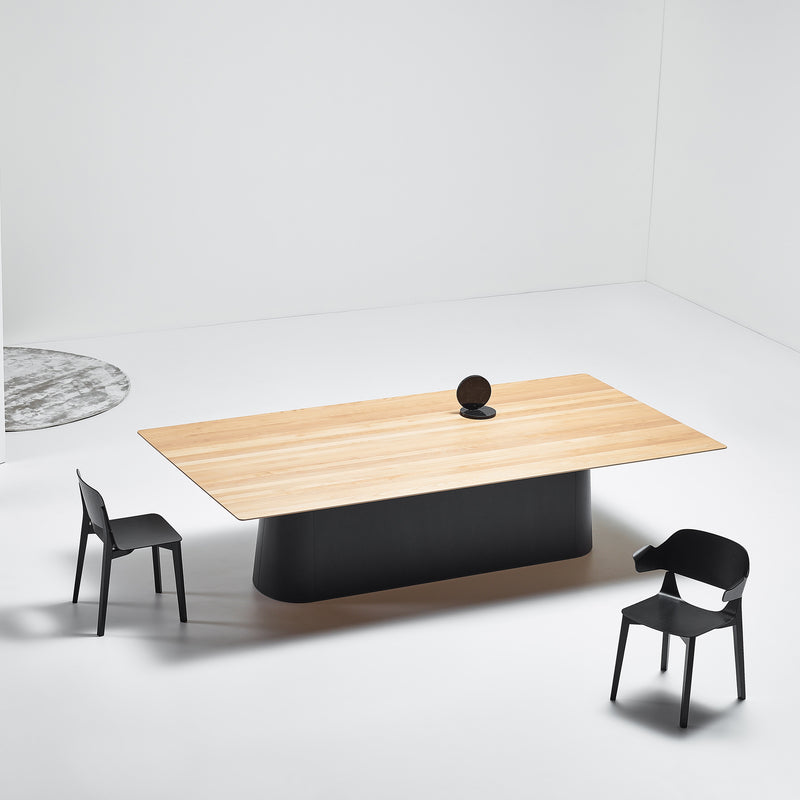 A minimalist room showcases a P.O.V. Table 467 by TON, featuring an organic black base and two black chairs. A small round object adorns the table, while white walls and floors contrast with a glimpse of a round gray rug in the corner.