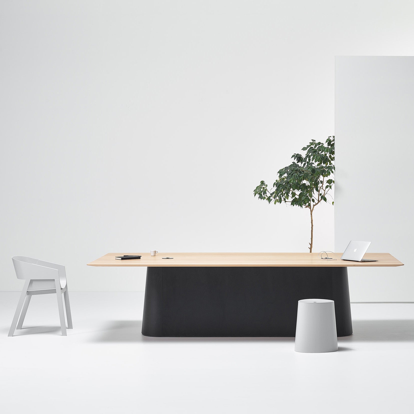 A minimalist office setup showcases the sleek P.O.V. Table 467 by TON, adorned with a laptop, notepad, and small plant. Nearby, a modern chair and cylindrical stool sit on the white floor. A larger potted plant adds greenery to the backdrop, emphasizing the modular space design.
