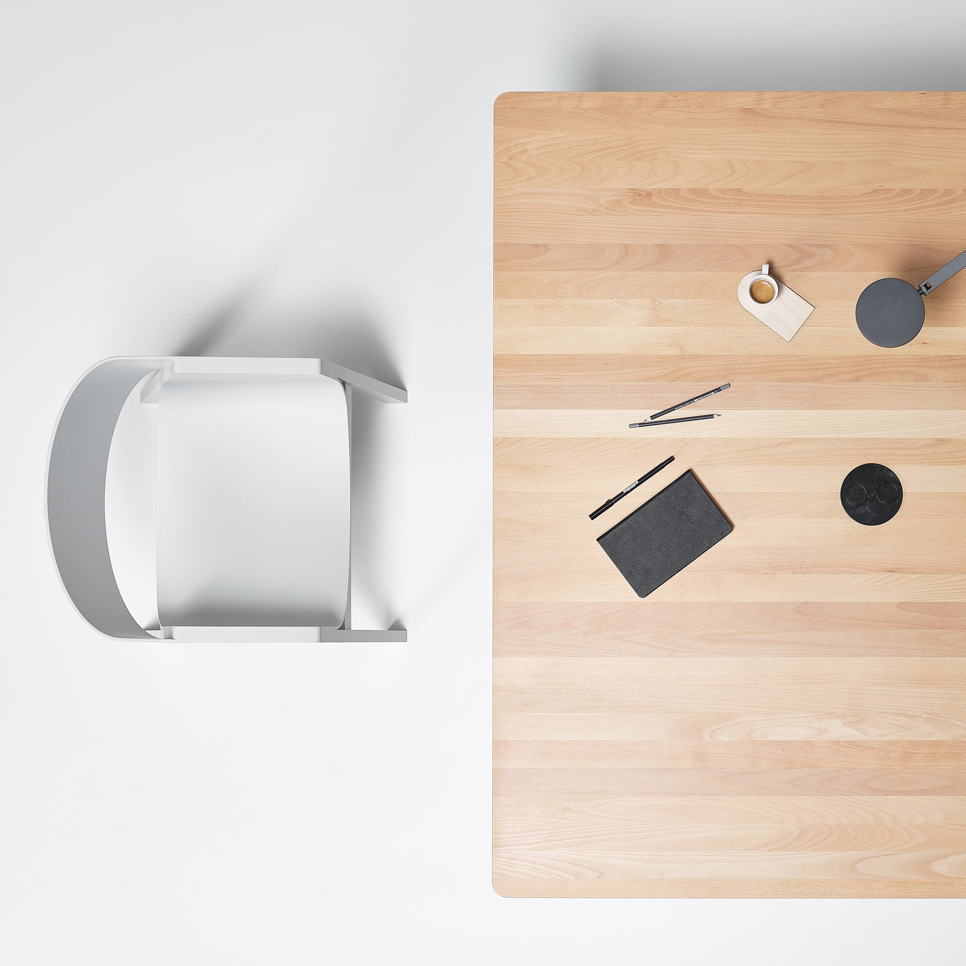 Minimalist workspace aerial view featuring a P.O.V. Table 467 by TON and a white chair. On the table are a closed black notebook, pens, coffee on an organic table base coaster, and a cardboard tag against a plain white surface.