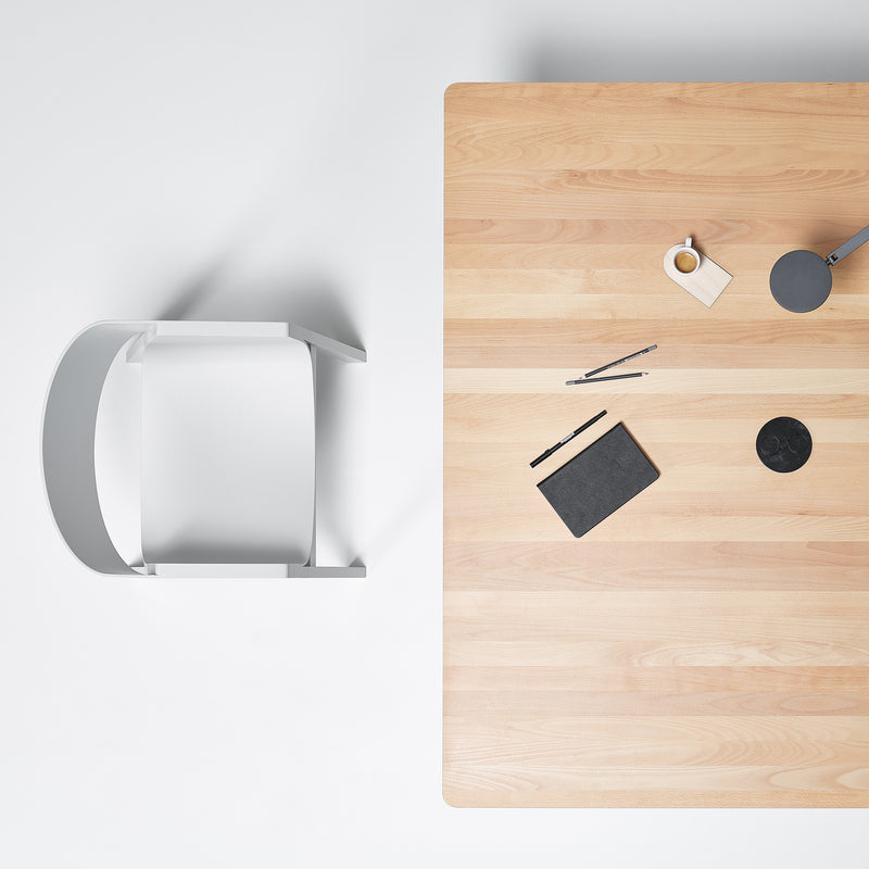 Minimalist workspace aerial view featuring a P.O.V. Table 467 by TON and a white chair. On the table are a closed black notebook, pens, coffee on an organic table base coaster, and a cardboard tag against a plain white surface.