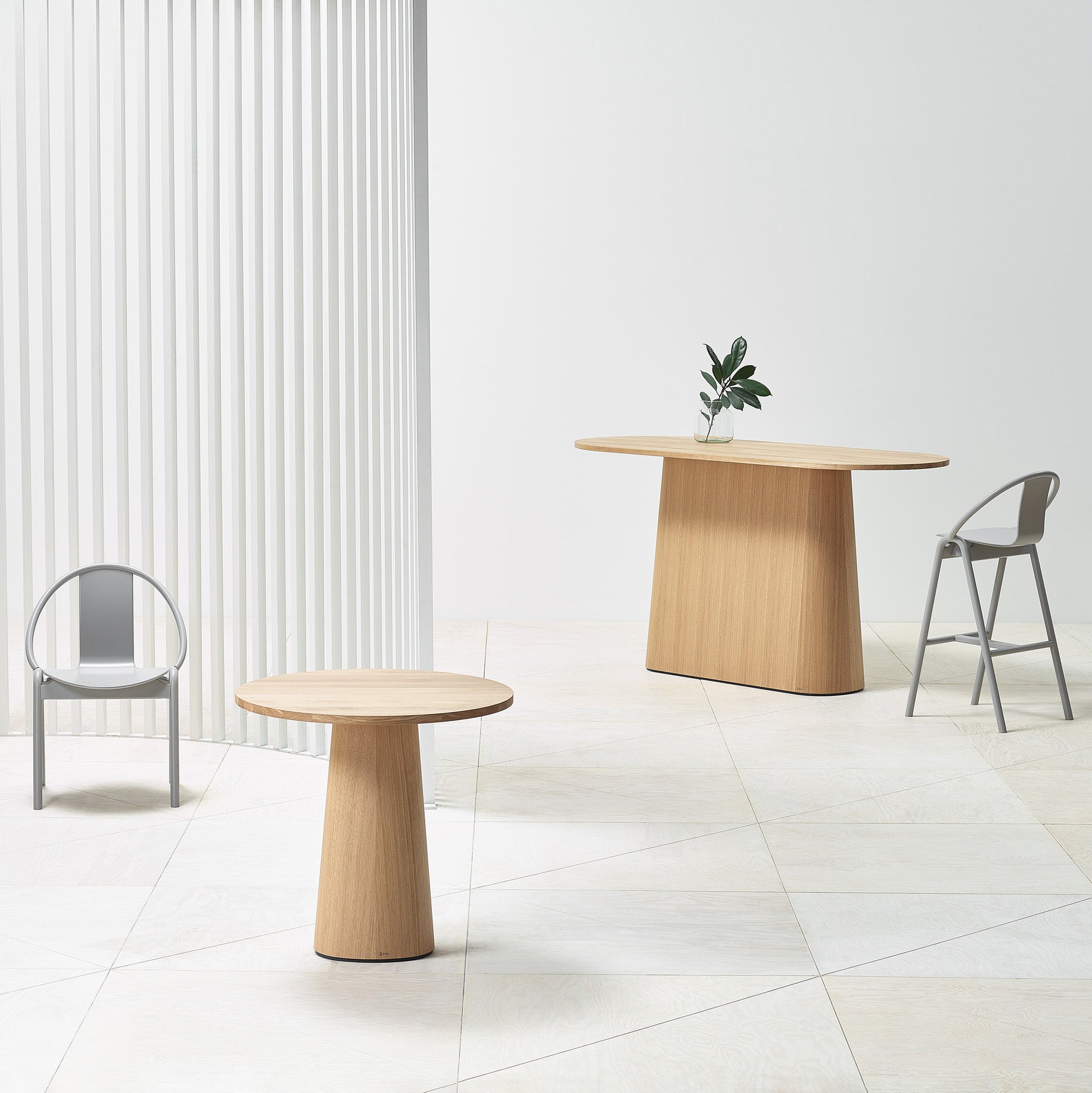 A minimalist room features the P.O.V. Table 468 by TON, a round wooden piece with an organic base, and chairs on a light tiled floor. Nearby, a larger wooden table with a plant complements the white curtain, creating a bright and modern setting.