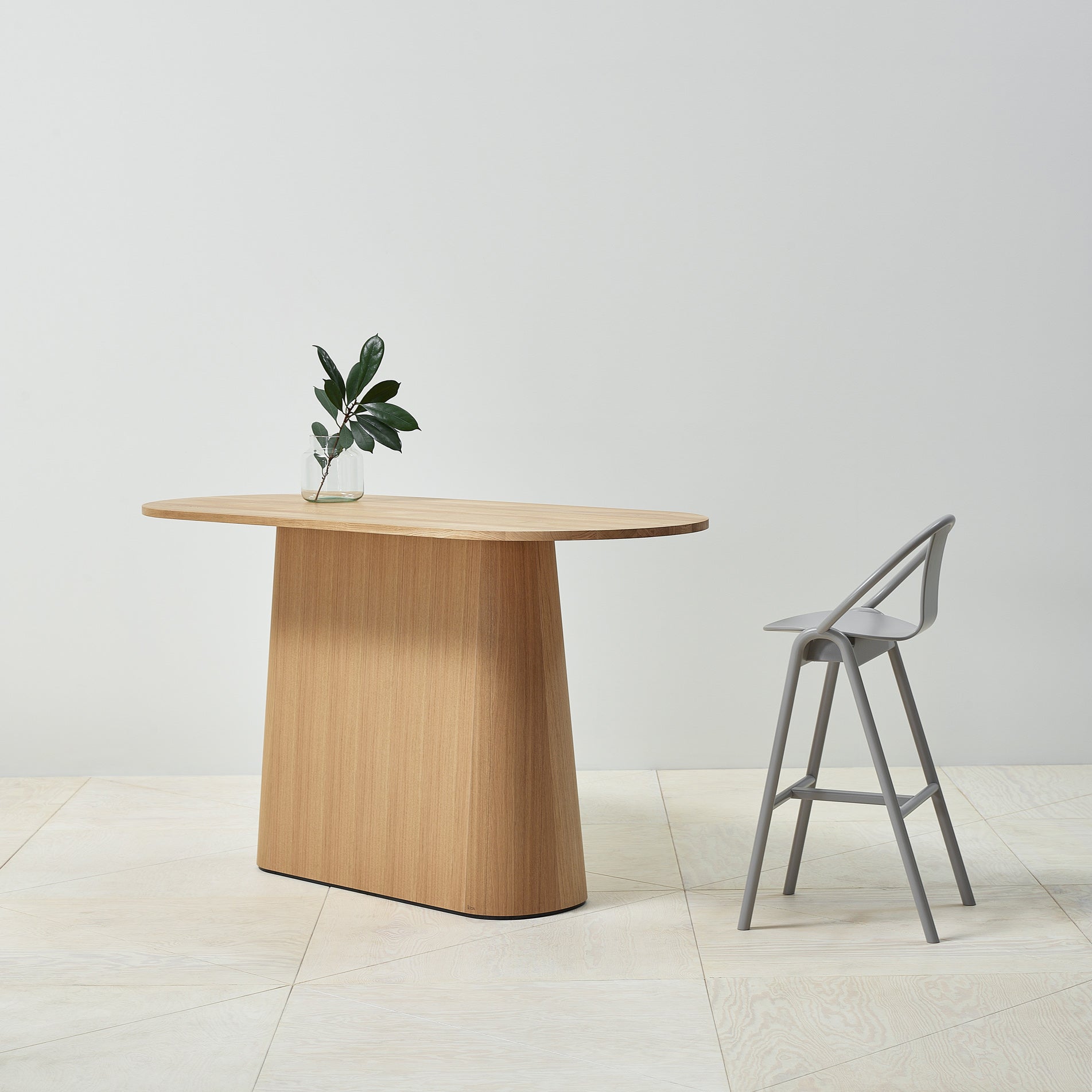 A modern, minimalist scene showcases the P.O.V. Table 468 by TON with an organically shaped base and a vase of green leaves on top. A sleek gray chair complements the table, all set against a plain white background on a light wood floor.