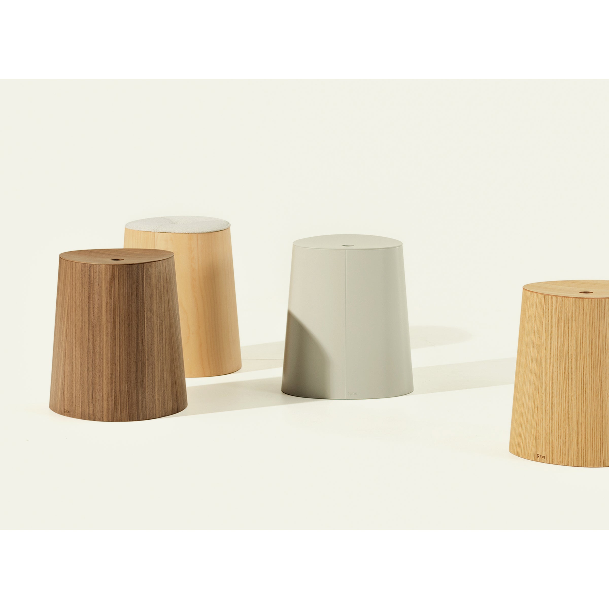 The P.O.V. Stool Upholstered by TON showcases four stackable dining stools in shades of brown and gray, crafted from American walnut. These stools feature a simple cylindrical design with slightly tapered sides, arranged on a light background with soft lighting.