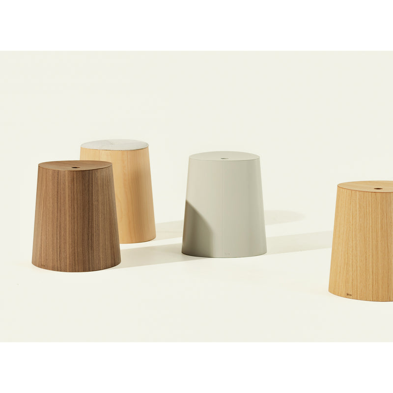 The P.O.V. Stool Upholstered by TON showcases four stackable dining stools in shades of brown and gray, crafted from American walnut. These stools feature a simple cylindrical design with slightly tapered sides, arranged on a light background with soft lighting.