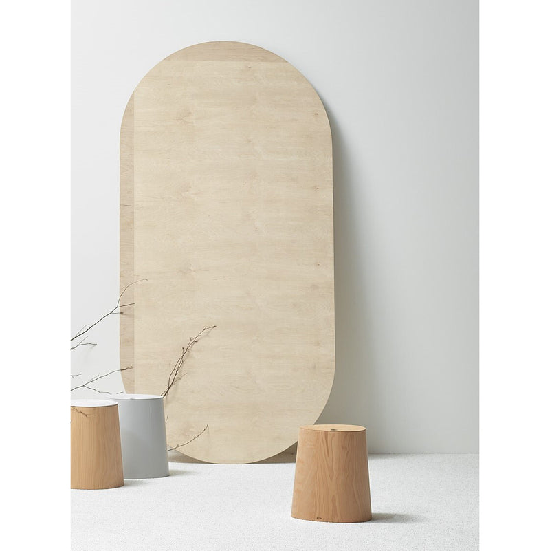 In a minimalist setting, a large wooden oval panel leans against a light wall. From TONs P.O.V. collection, three cylindrical wooden stools and one gray metal stool are in front, with twigs on the ground adding natural charm to the serene scene.