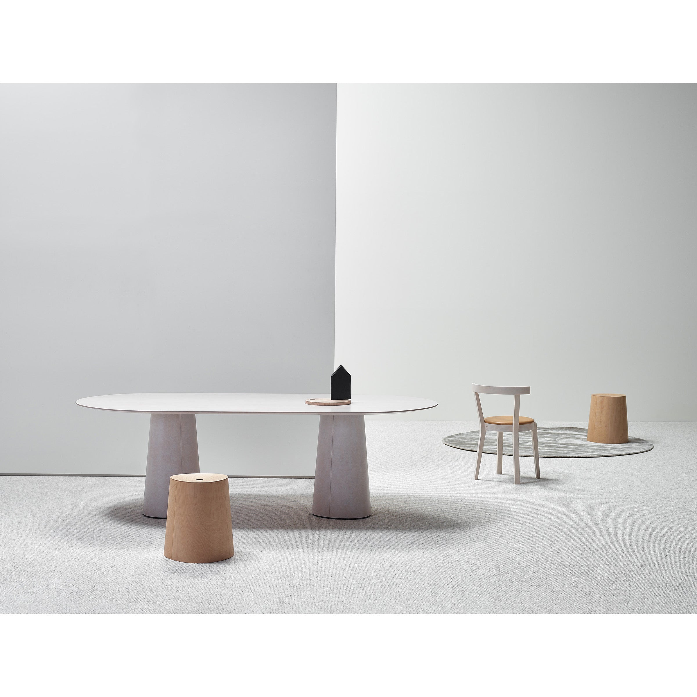 A minimalist interior features a large oval table with two American walnut cylindrical stools, a small black house-shaped decor, and the P.O.V. Stool Upholstered by TON. A white chair and round gray rug complete the scene set against white walls and carpeting.