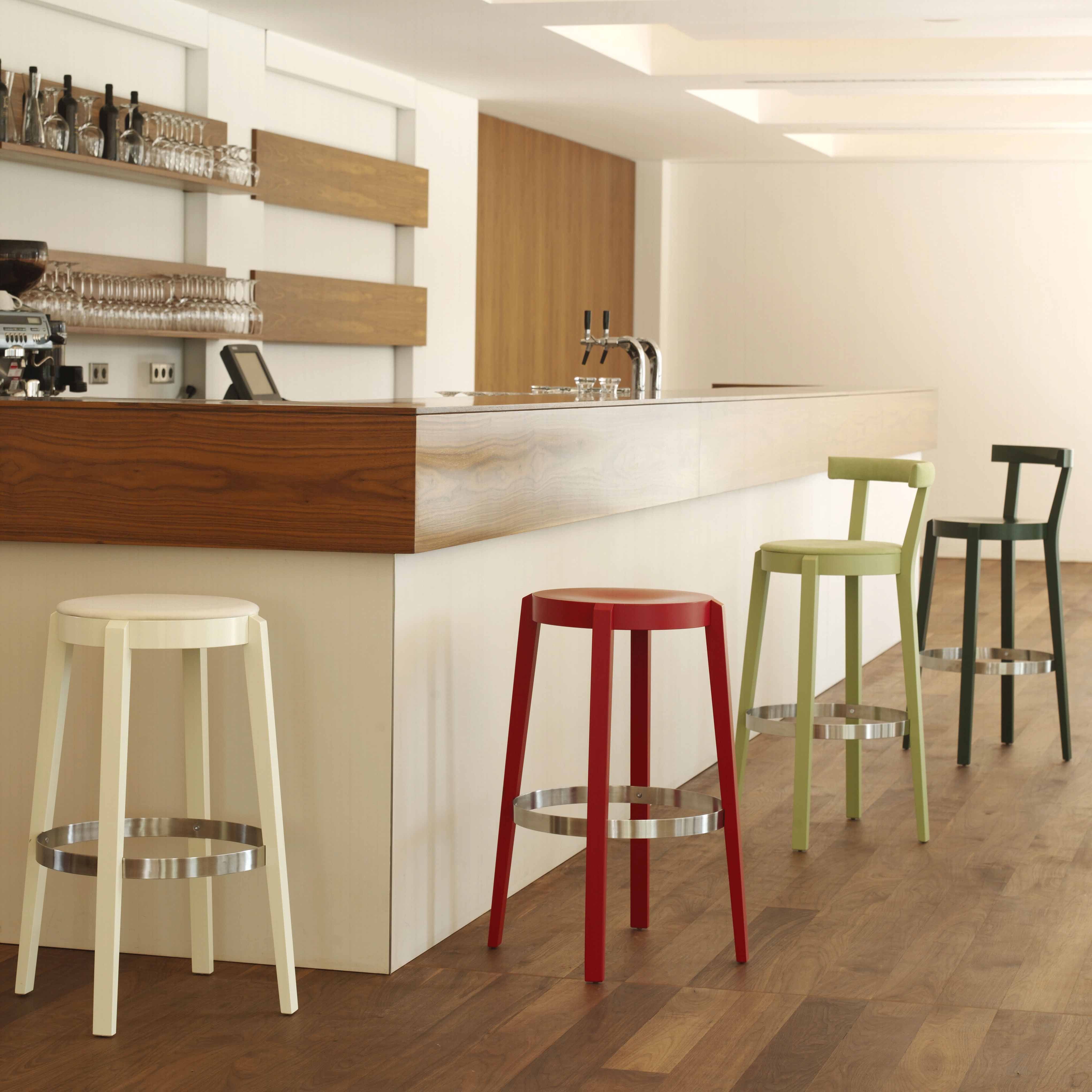 A modern bar area showcases a sleek wooden counter and floor. Arranged neatly along the bar are three TON Barstool Punton chairs, each with beech legs in cream, red, and green. Behind them, shelves of glassware and coffee equipment enhance the clean design.